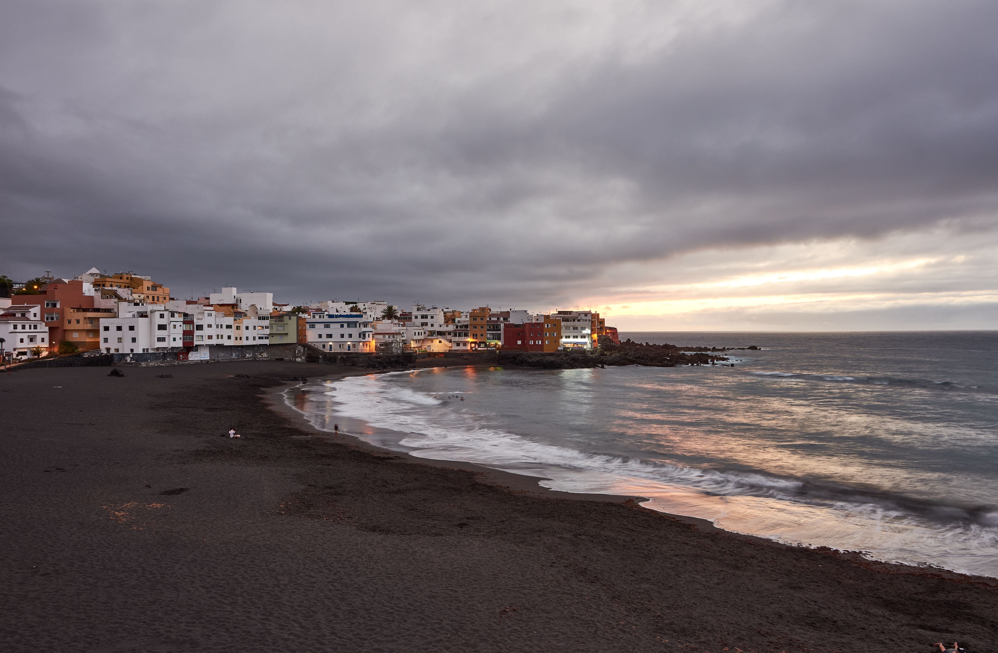 Dunkler Sandstrand bei Sonnenuntergang mit bunten Häusern in Puerto de la Cruz, Teneriffa.
