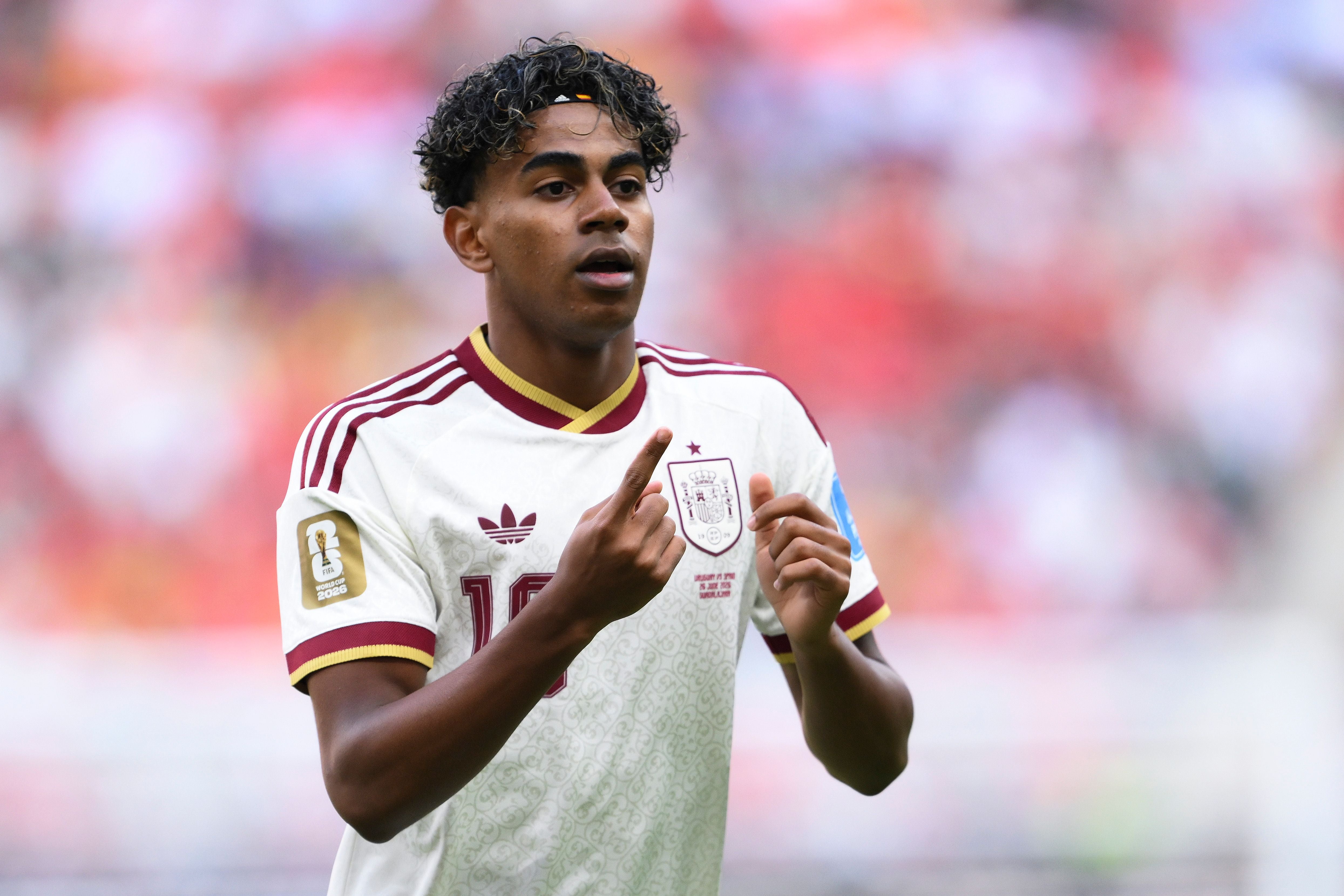 Ein spanischer Fußballspieler im weißen Trikot zeigt auf das Spanien-Wappen auf seiner Brust.