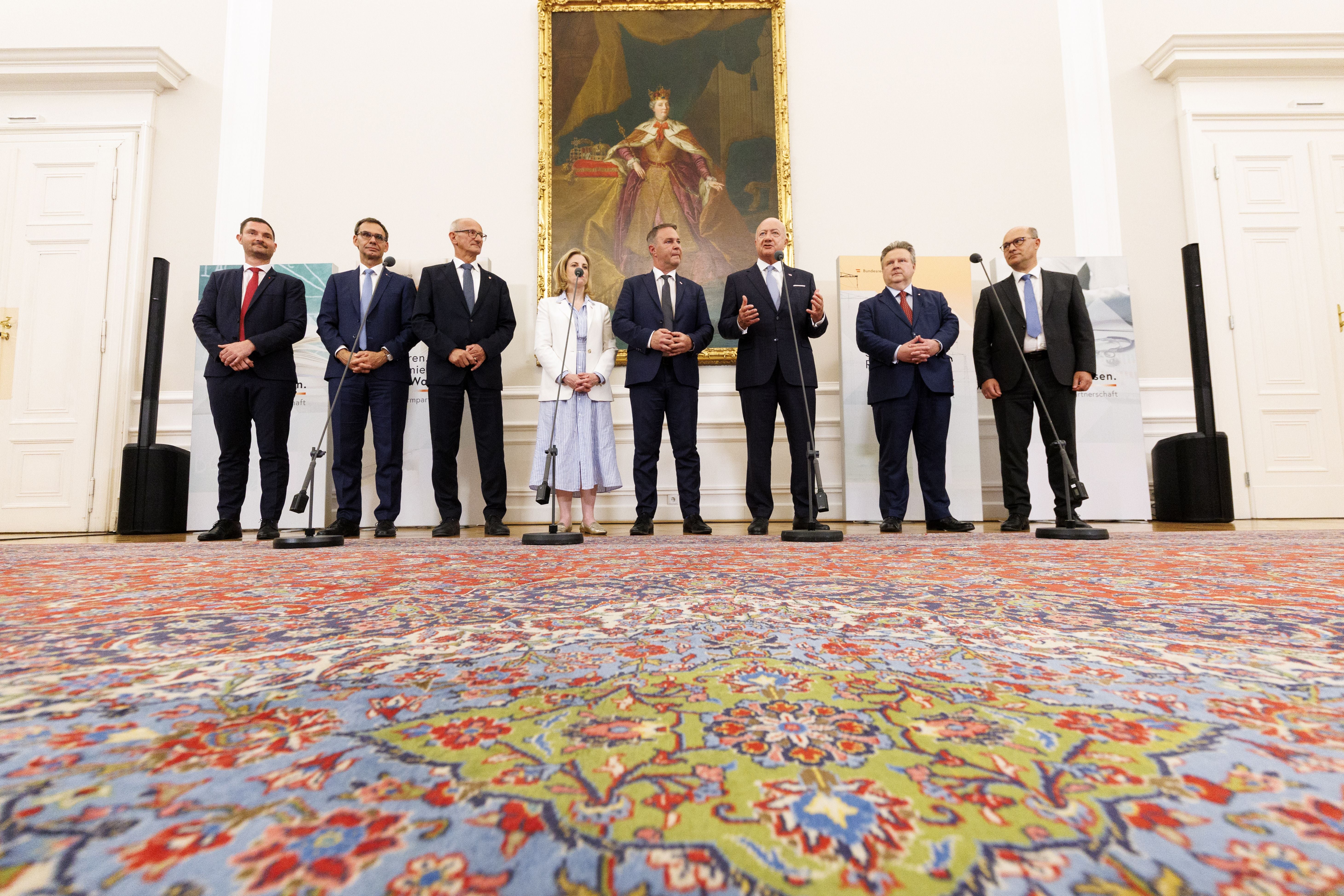 Acht Personen stehen bei einer Pressekonferenz im Bundeskanzleramt Wien auf einem bunten Teppich.