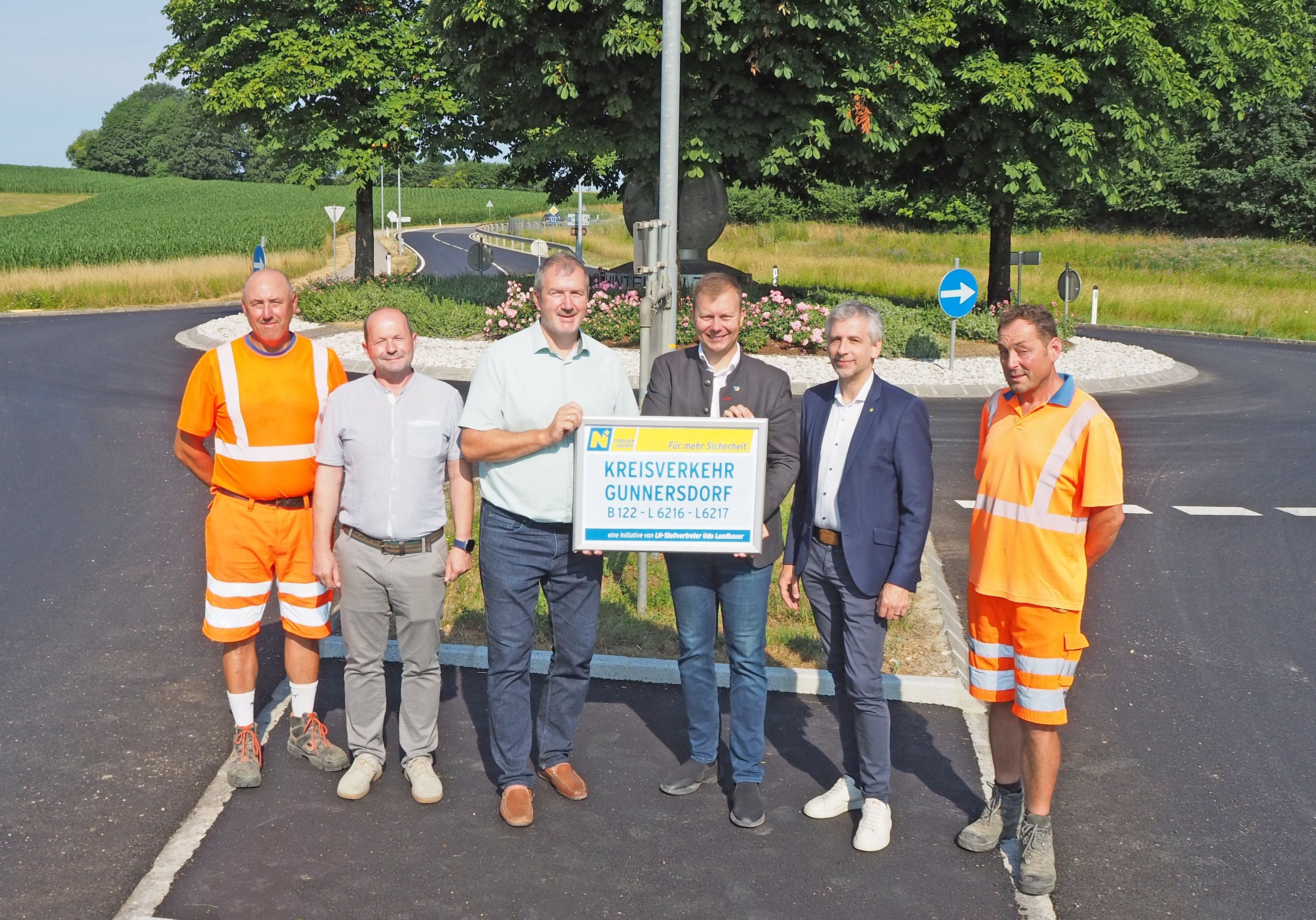 Sechs Männer stehen vor einem Kreisverkehr, zwei tragen orange Arbeitskleidung, einige halten ein Schild.