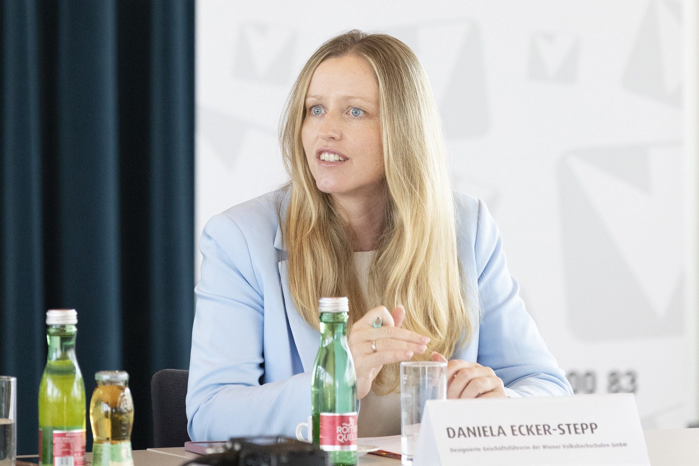 Frau mit langen blonden Haaren spricht bei einer Konferenz, vor sich Wasserflaschen und Namensschild.