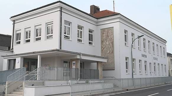 Modernes, weißes Schulgebäude mit vielen Fenstern und einer Reliefwand an der Fassade.