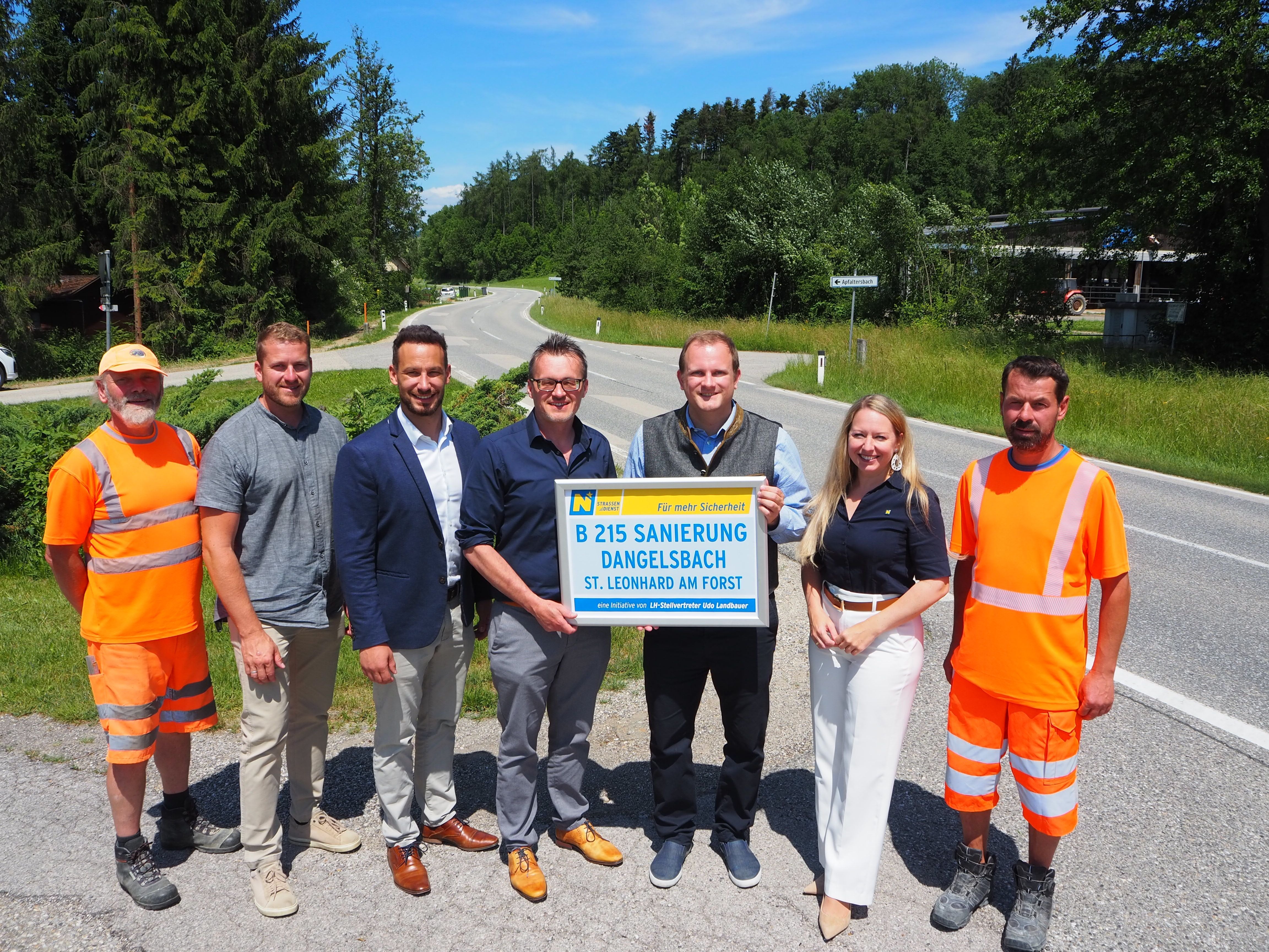 Acht Personen stehen an einer Straße und halten ein Schild zur Straßensanierung hoch.