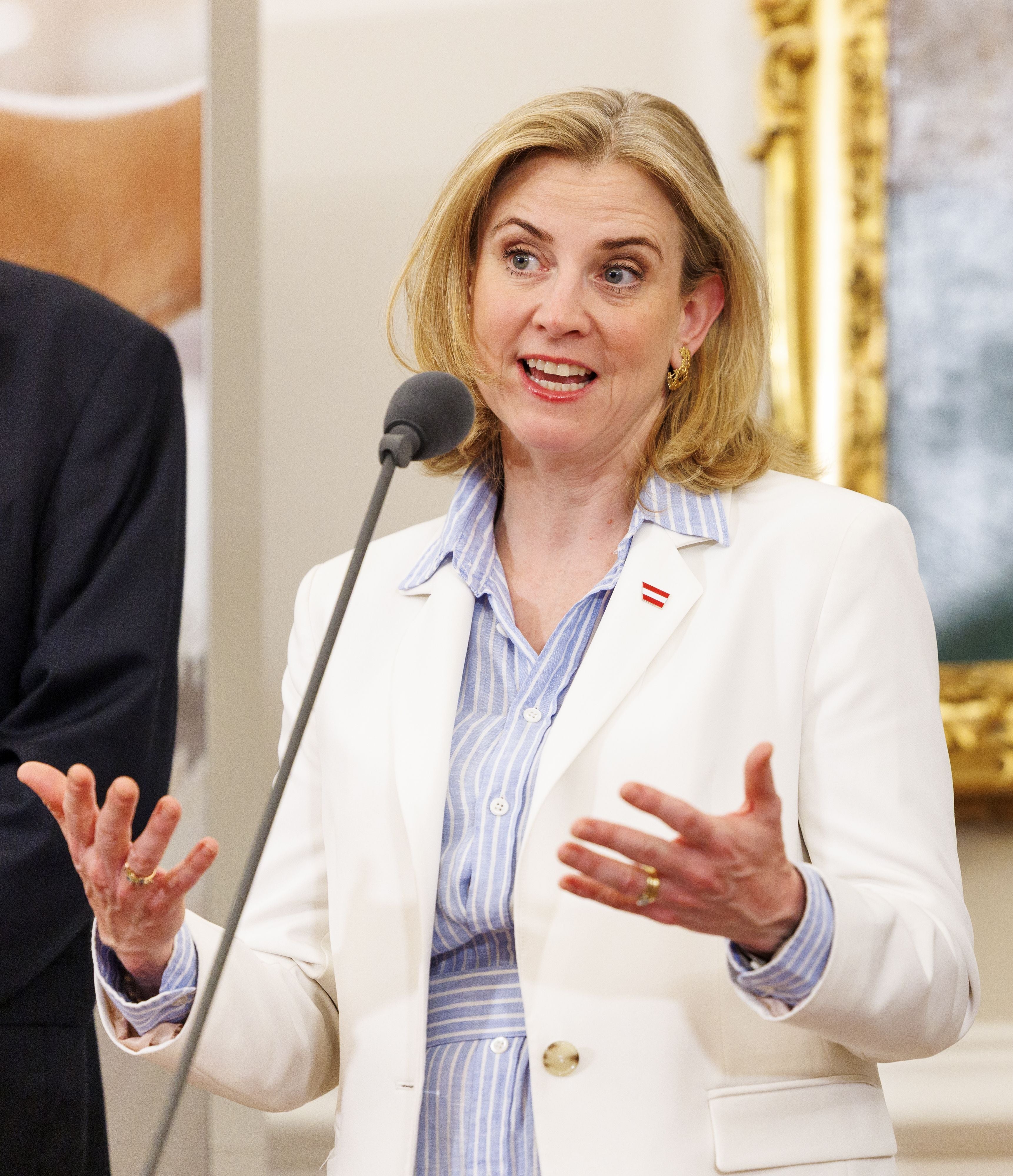 Eine Frau im weißen Blazer spricht gestikulierend bei einer Pressekonferenz am Mikrofon.
