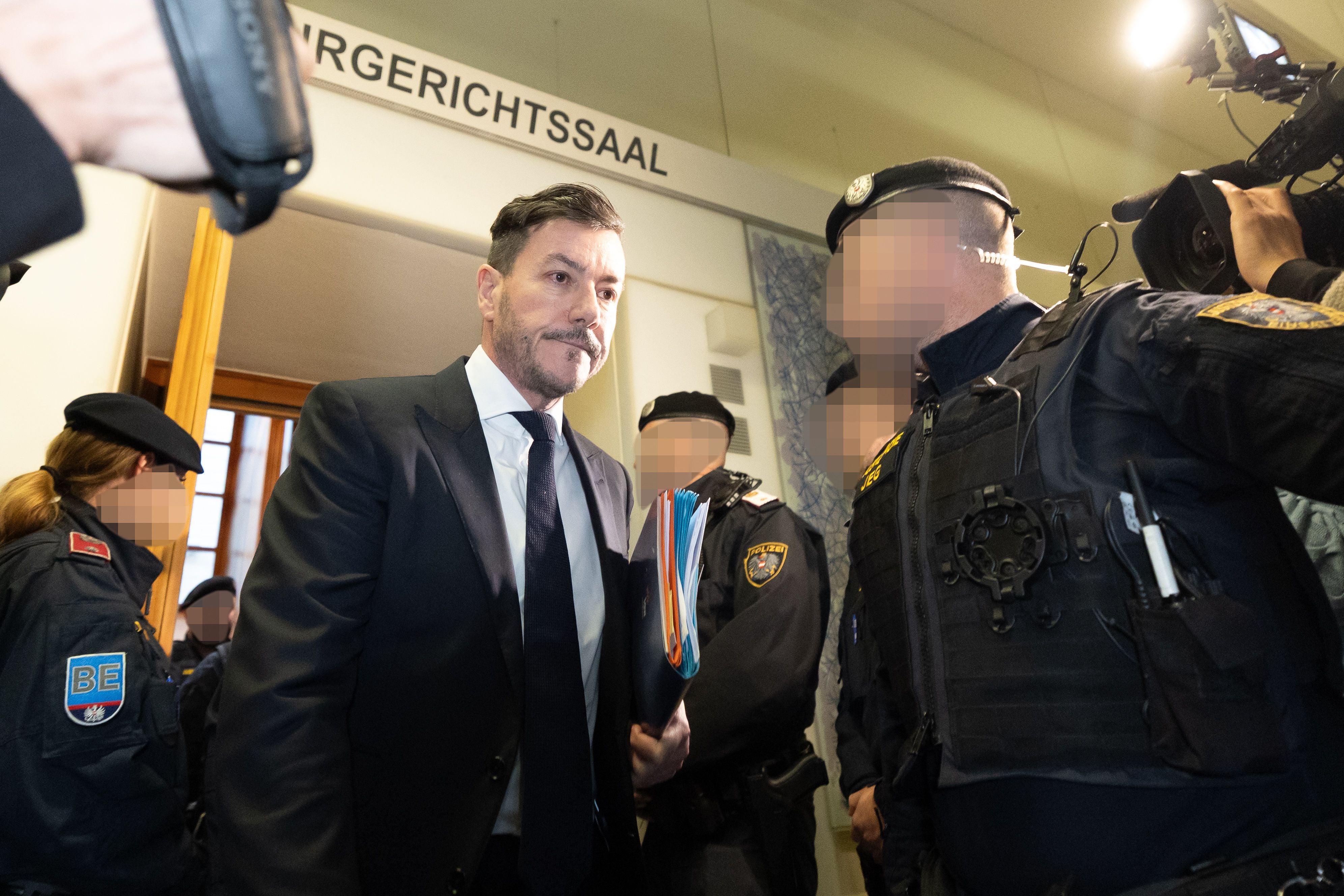 Mehrere Polizisten begleiten einen Anzugträger mit Akte in einen Gerichtssaal.