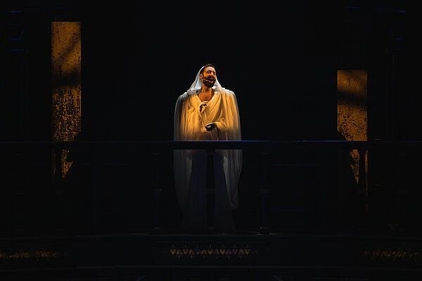 Eine Person im weißen Umhang steht auf einer dunklen Theaterbühne im Scheinwerferlicht.