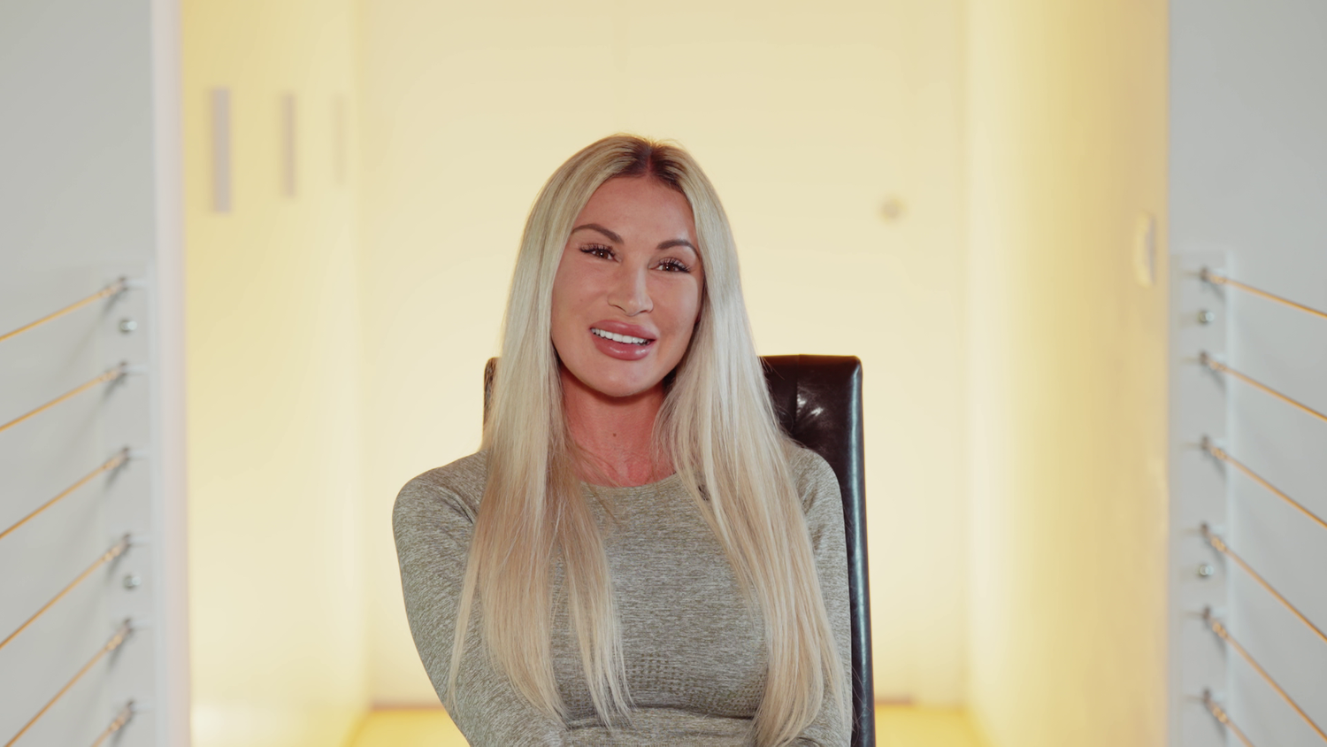 Frau mit langen blonden Haaren sitzt lächelnd auf einem Stuhl in einem hellen Raum.