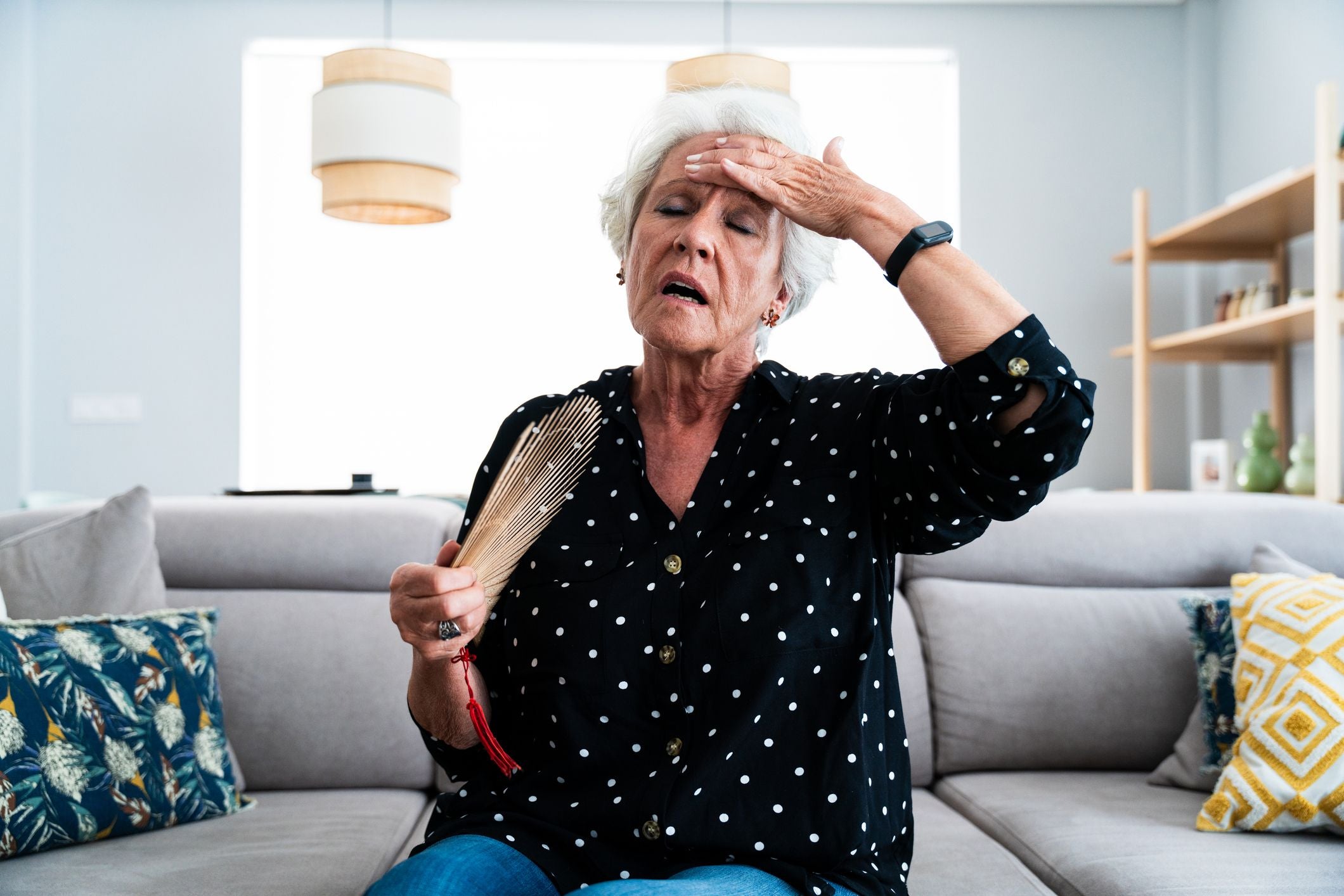 Ältere Frau sitzt auf dem Sofa, fächert sich Luft zu und wirkt erschöpft wegen Hitze.