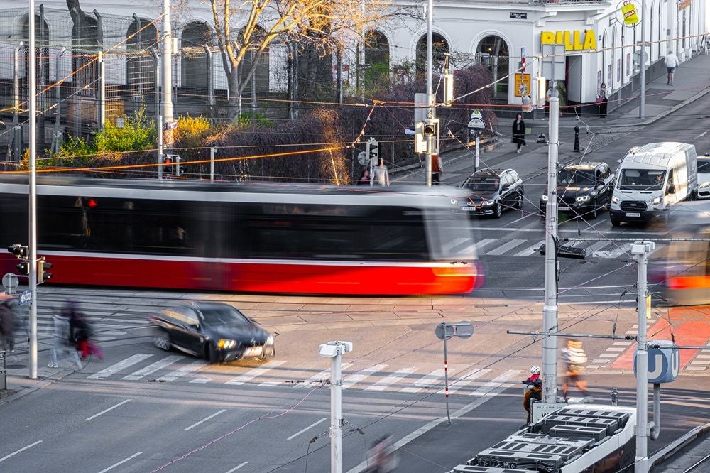 Straßenbahn fährt durch belebte Kreuzung, Autos und Passanten sind in Bewegung zu sehen.