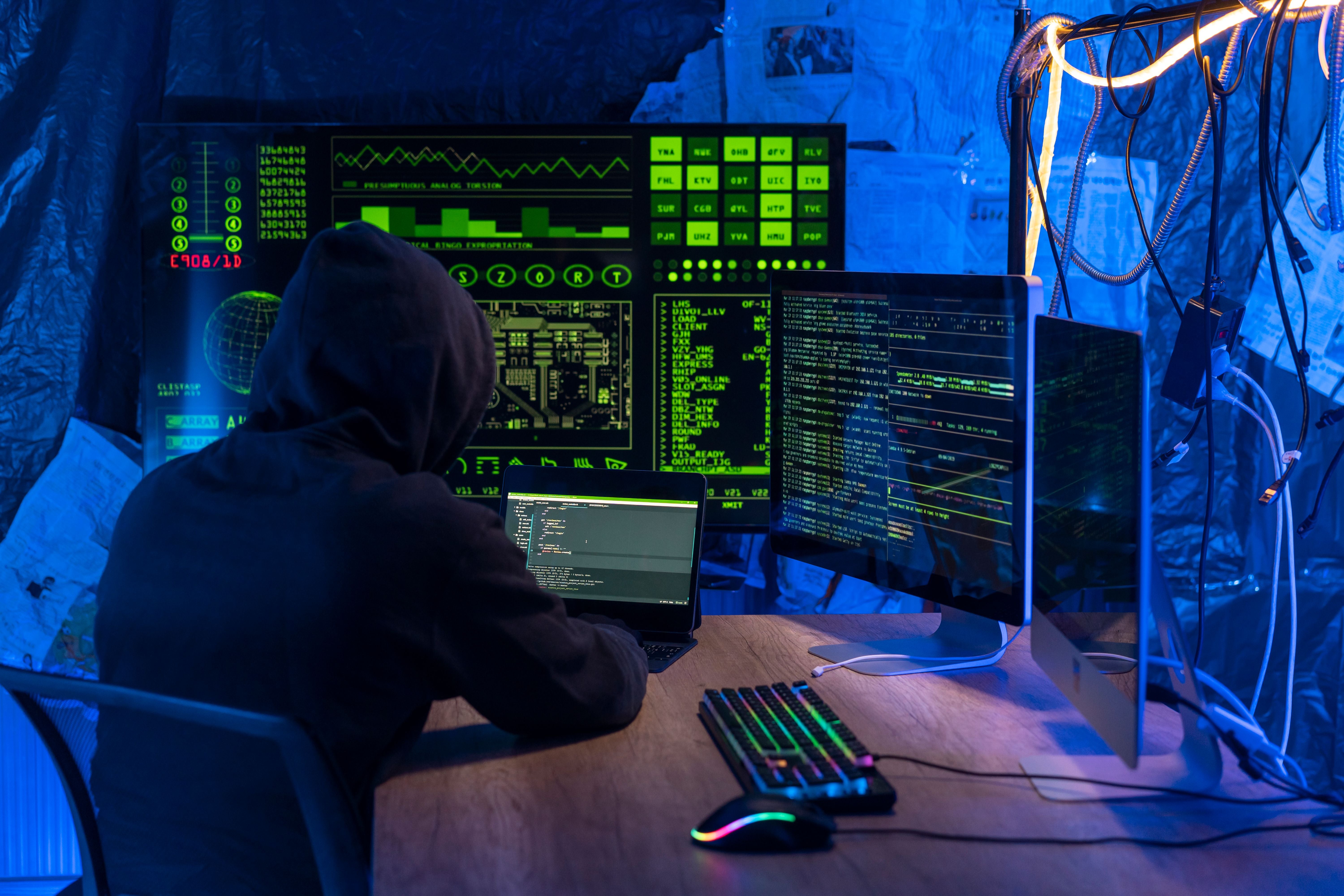 Eine Person in einem dunklen Raum mit Kapuzenpullover sitzt vor mehreren Monitoren mit grüner Codeanzeige, arbeitet an einem Laptop und einem Desktop-Computer; Kabel und bunt beleuchtete Tastatur deuten auf eine Szene aus dem Bereich Cyberkriminalität hin.