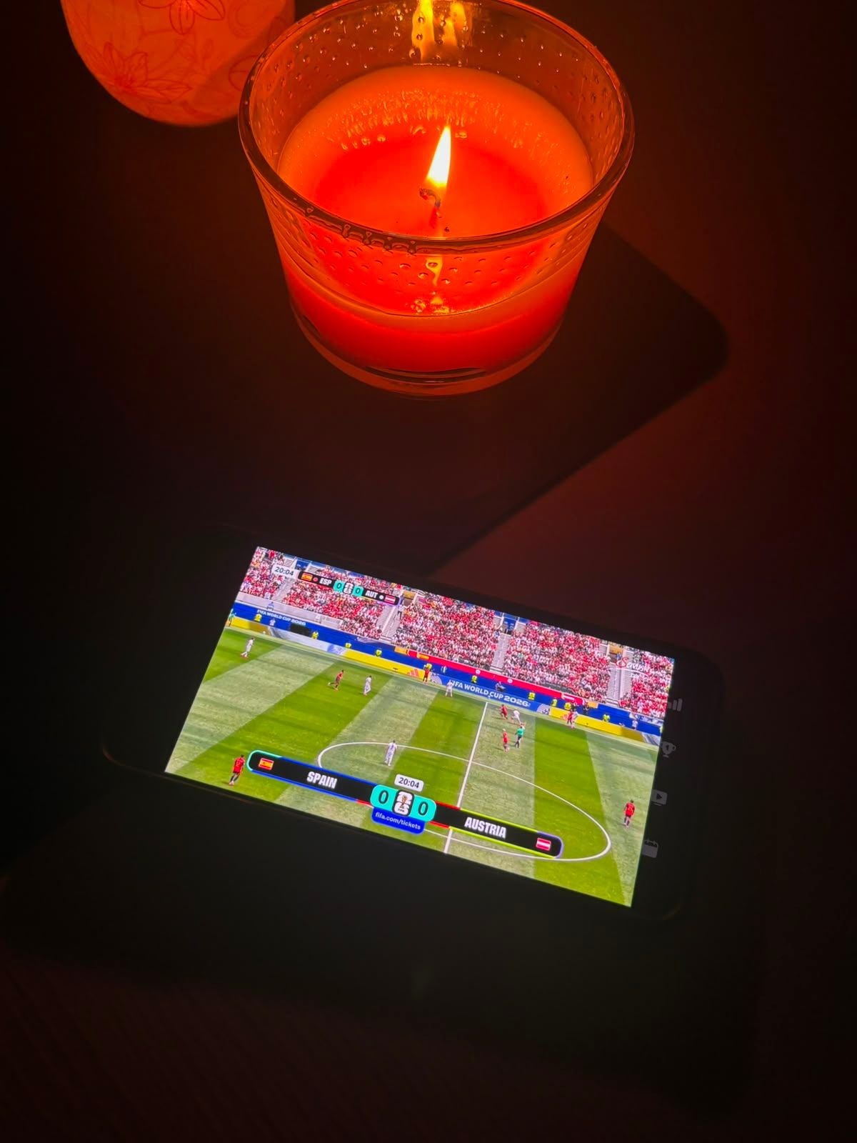 Ein brennende Kerze beleuchtet ein Handy, auf dem ein Fußballspiel läuft, vermutlich bei Stromausfall.