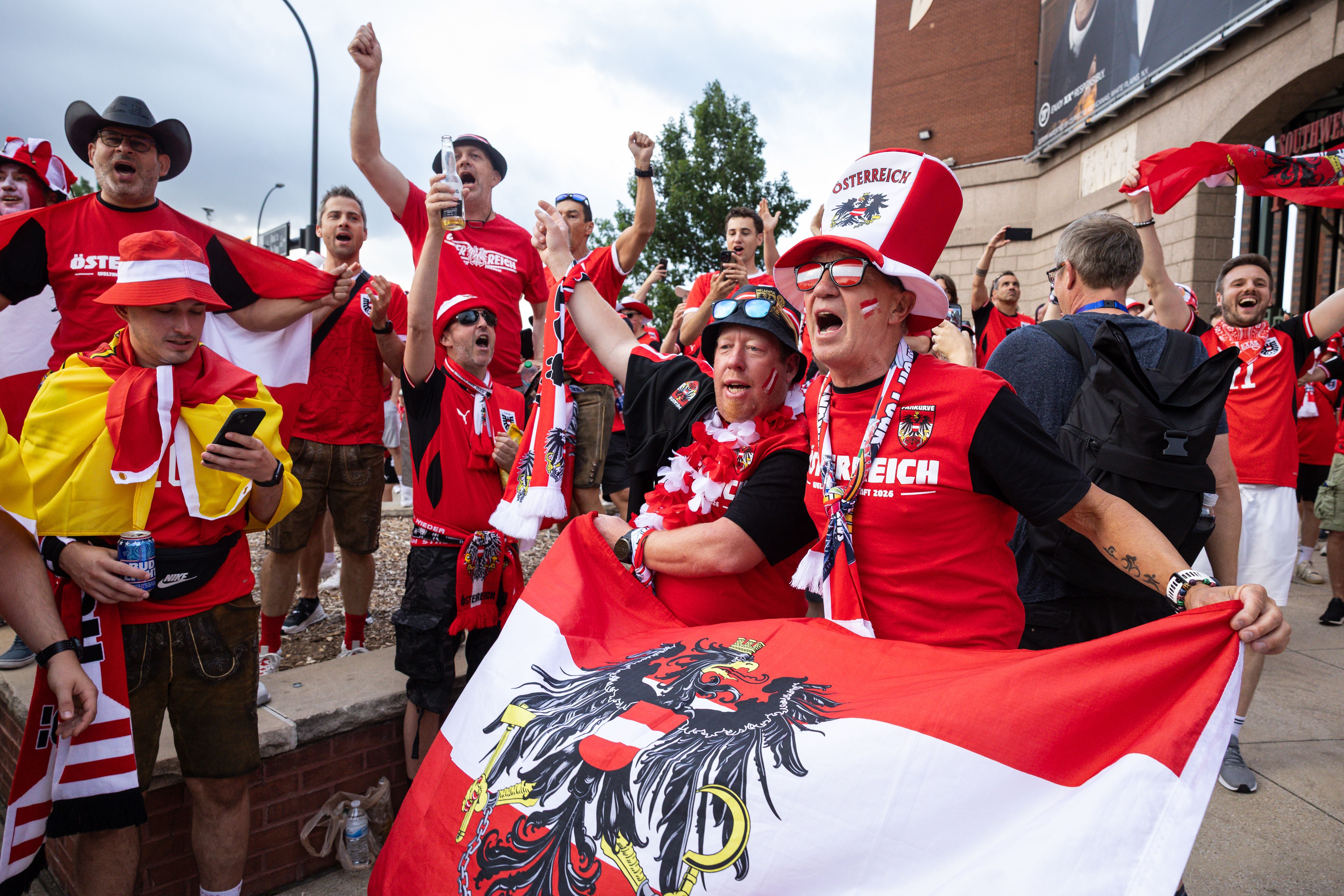 Österreichische Fußballfans jubeln in roten Shirts mit Flagge und Fanartikeln vor einem Stadion.