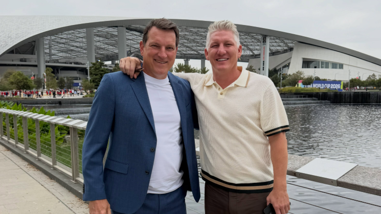 Zwei Männer stehen lächelnd vor einem Stadion an einem Gewässer.
