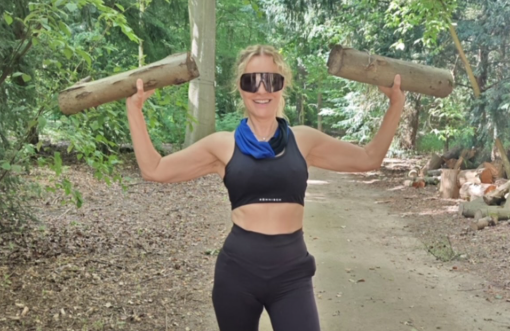 Frau im Sportoutfit hebt zwei Holzstücke im Wald und lächelt in die Kamera.
