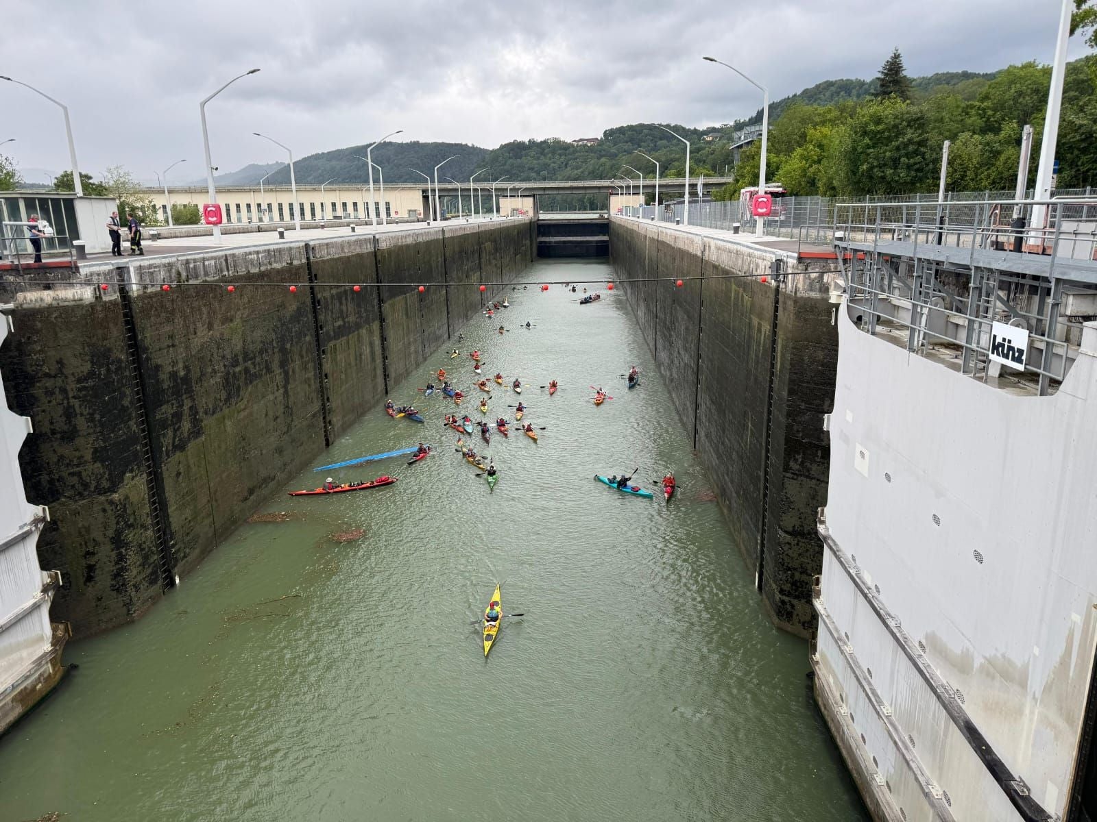 Mehrere Kanus mit Personen in einer Schleuse unter bewölktem Himmel und regnerischem Wetter.