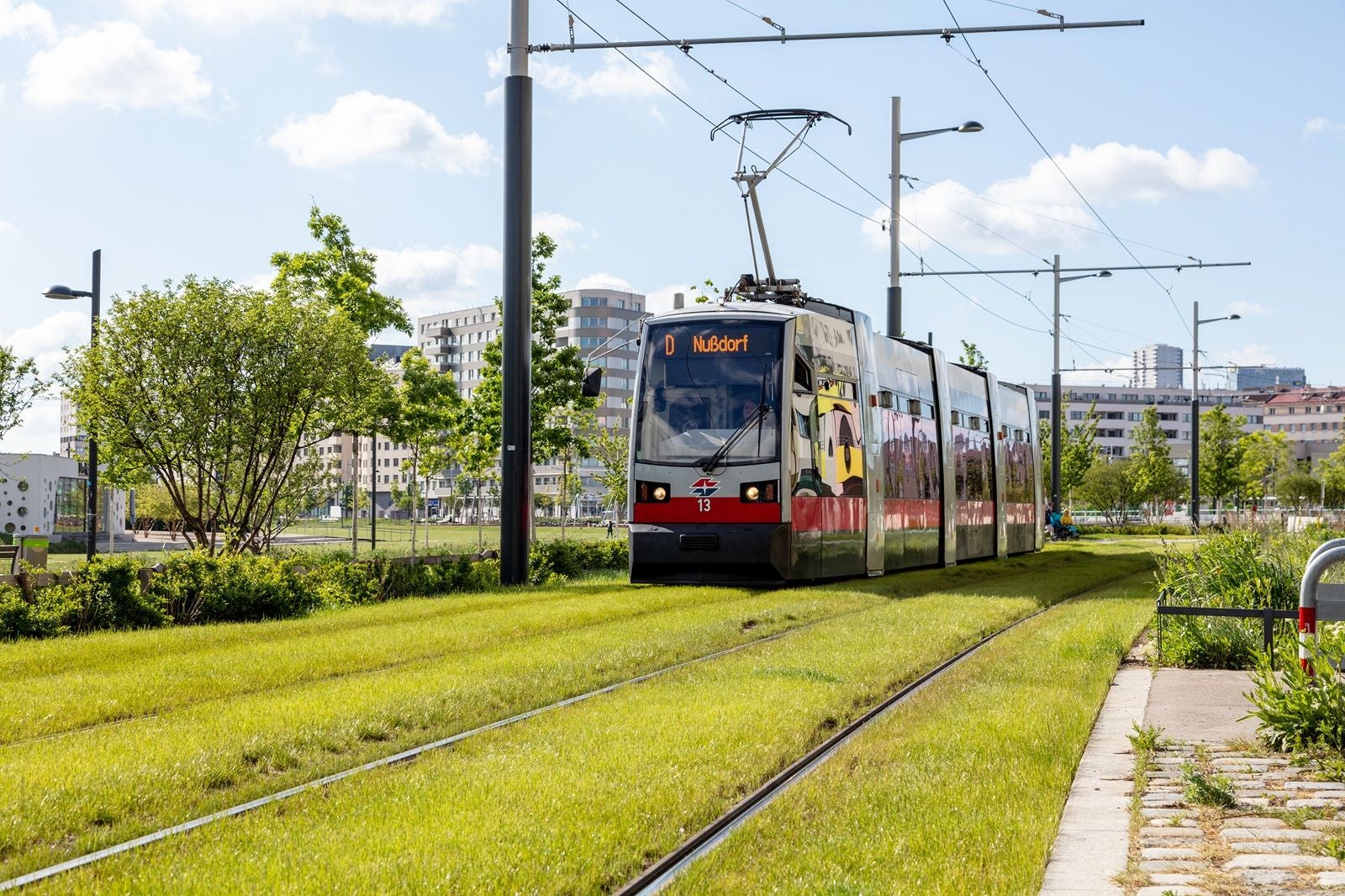 Straßenbahn fährt auf begrüntem Gleis durch eine Stadt mit modernen Gebäuden und Bäumen.