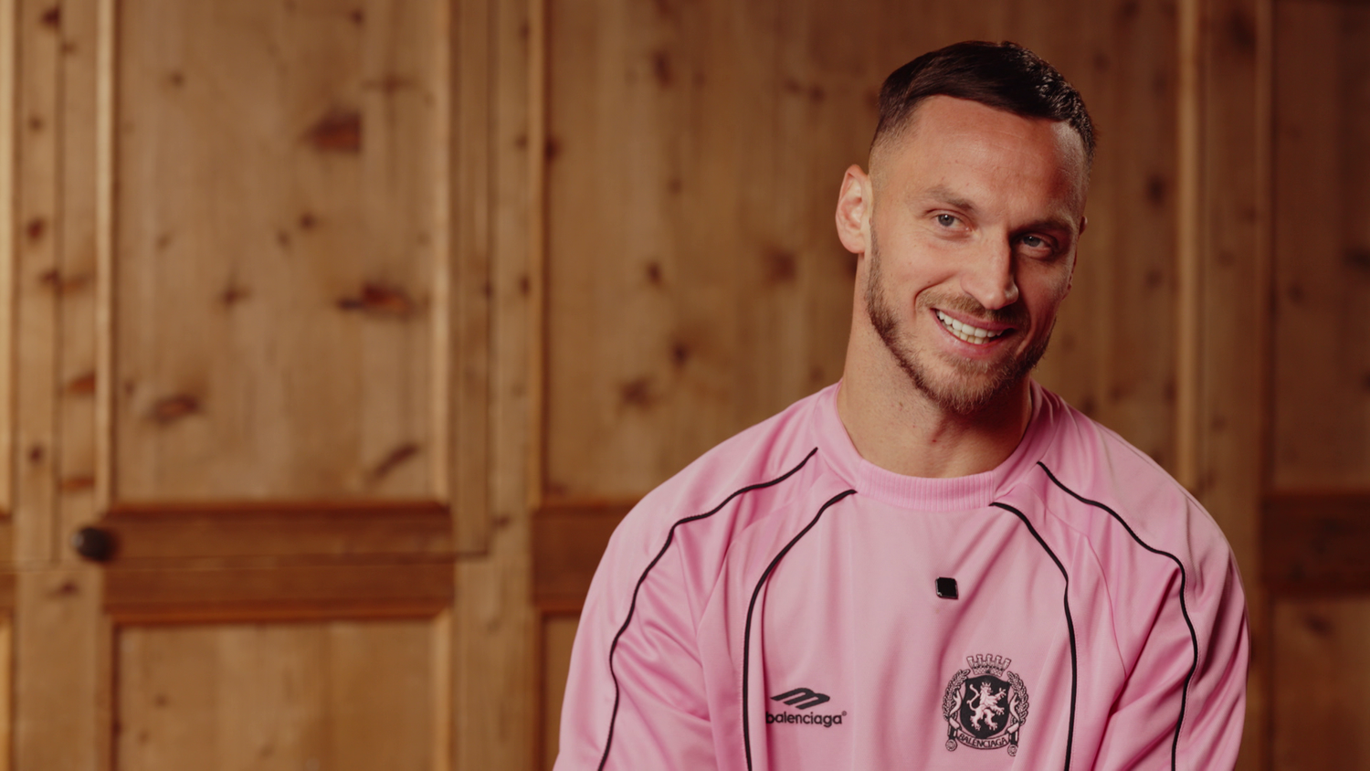 Ein Mann im rosa Trikot sitzt lächelnd vor einer hölzernen Wand.