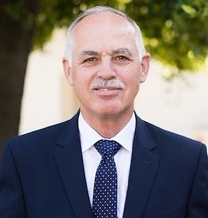 Ein älterer Mann im Anzug mit Krawatte steht im Freien und lächelt leicht in die Kamera.