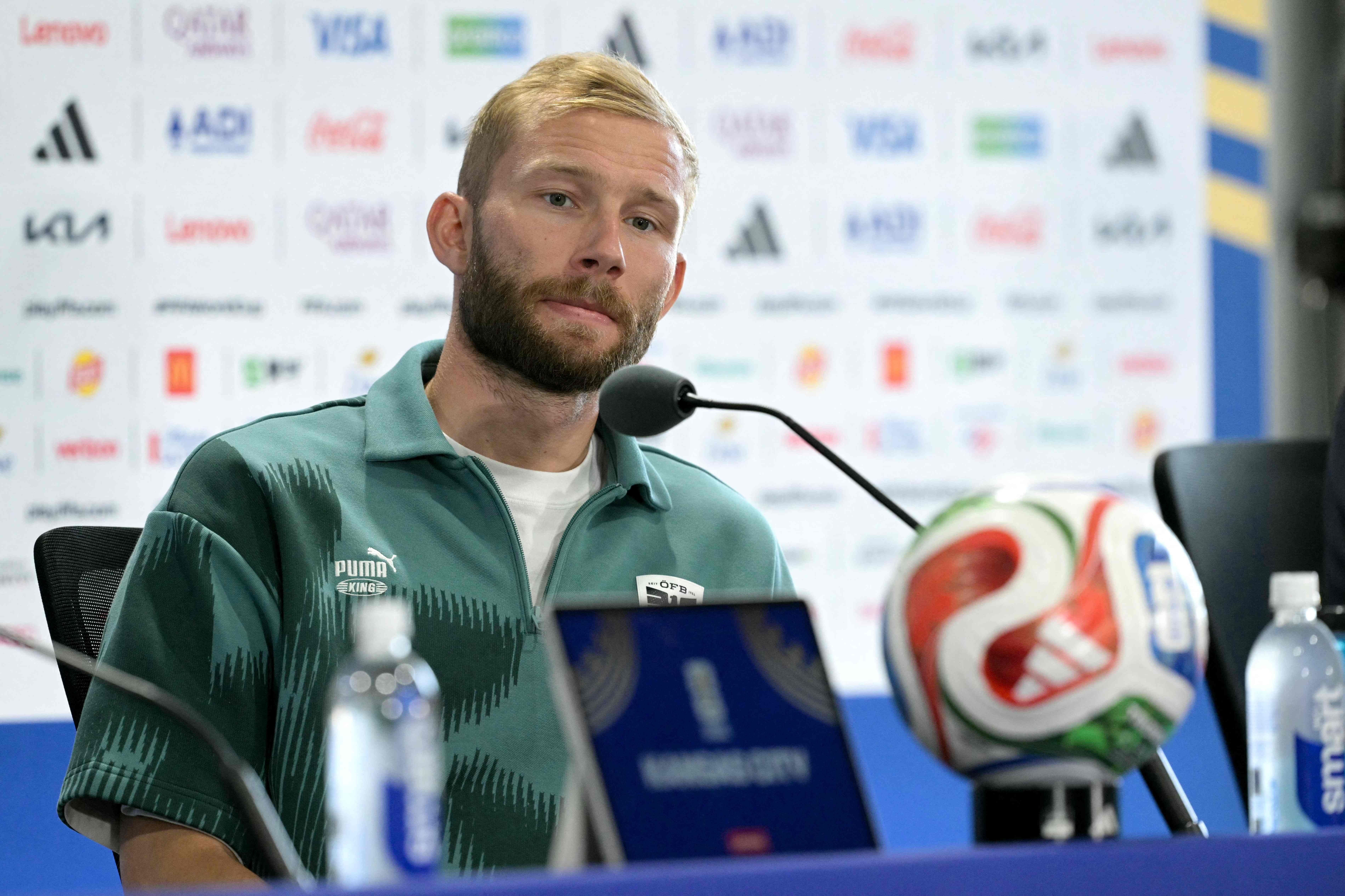 Ein Mann im grünen Puma-Pullover spricht auf einer Pressekonferenz vor Mikrofon und Fußball.