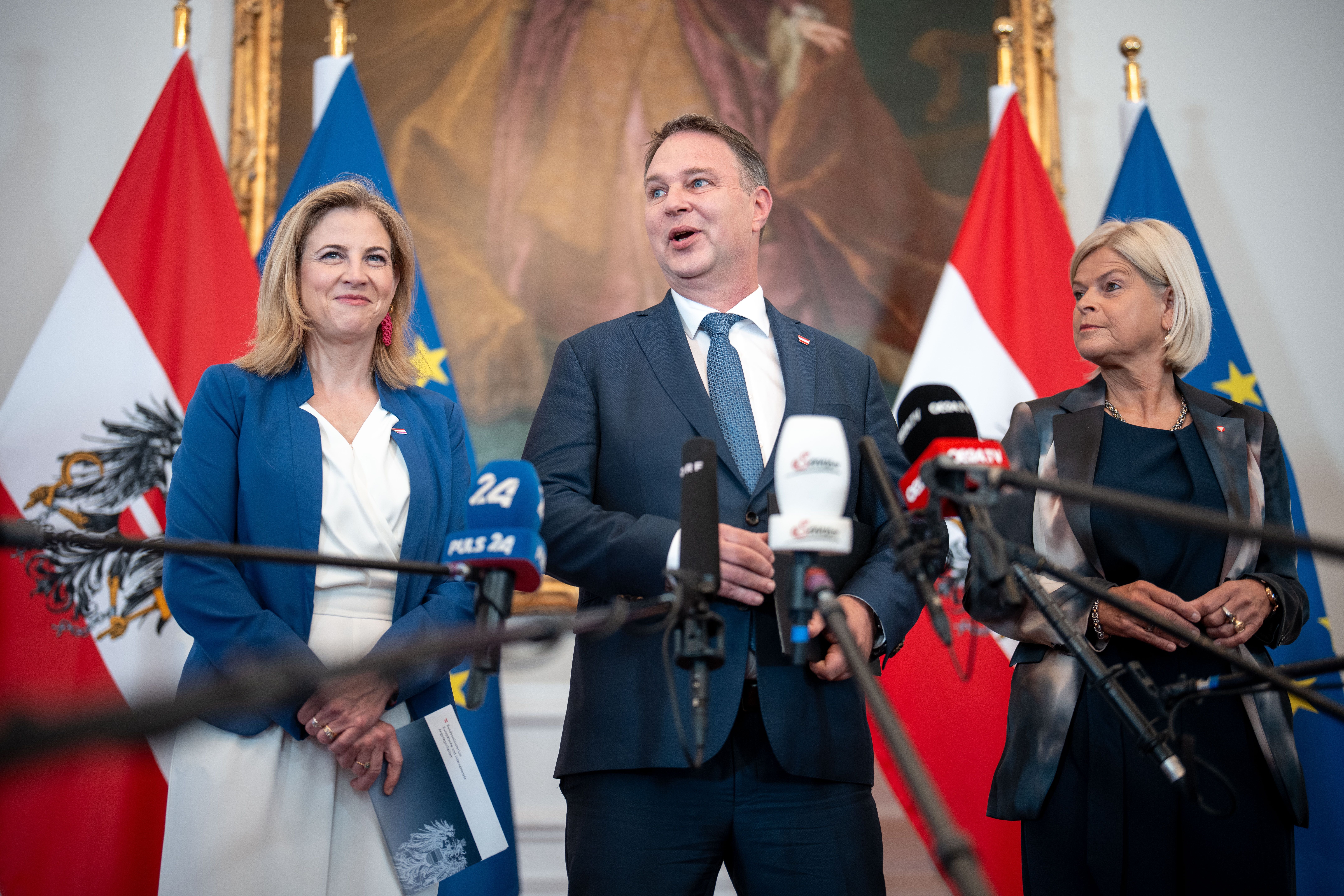 Drei Politiker bei einer Pressekonferenz im Bundeskanzleramt mit Mikrofonen und Flaggen im Hintergrund.