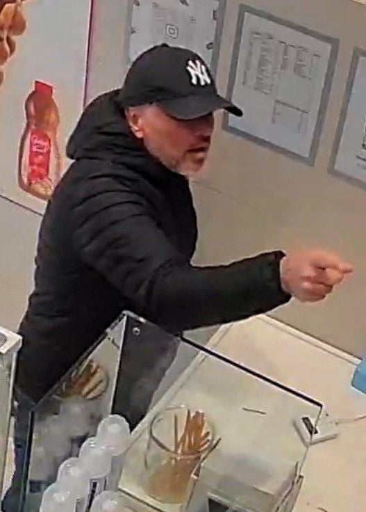 Ein Mann mit schwarzer Jacke und NY-Cap gestikuliert an einer Verkaufstheke.