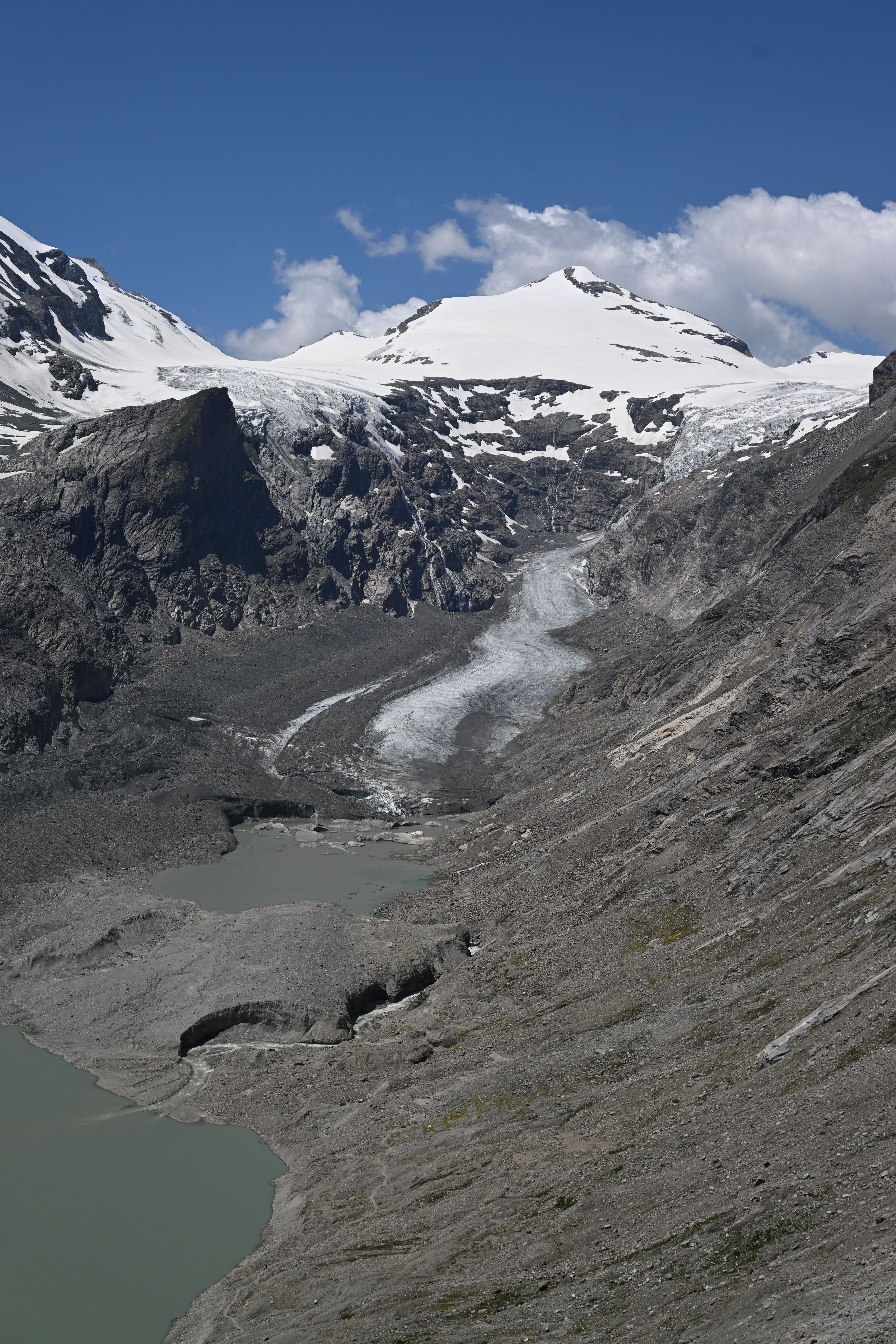 Blick auf die graue Pasterze, Gletschersee und schneebedeckte Berge unter blauem Himmel.