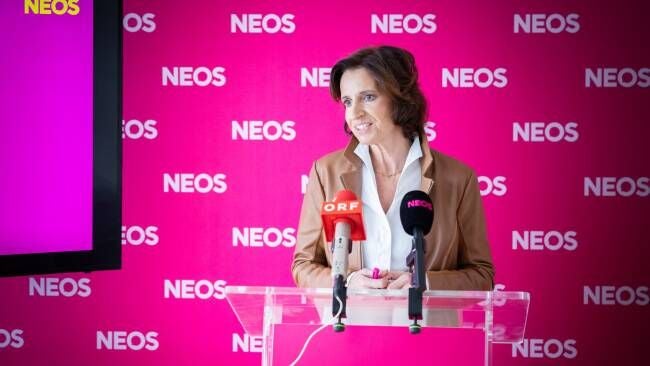 Frau spricht am Rednerpult vor pinkem NEOS-Hintergrund mit Mikrofonen.
