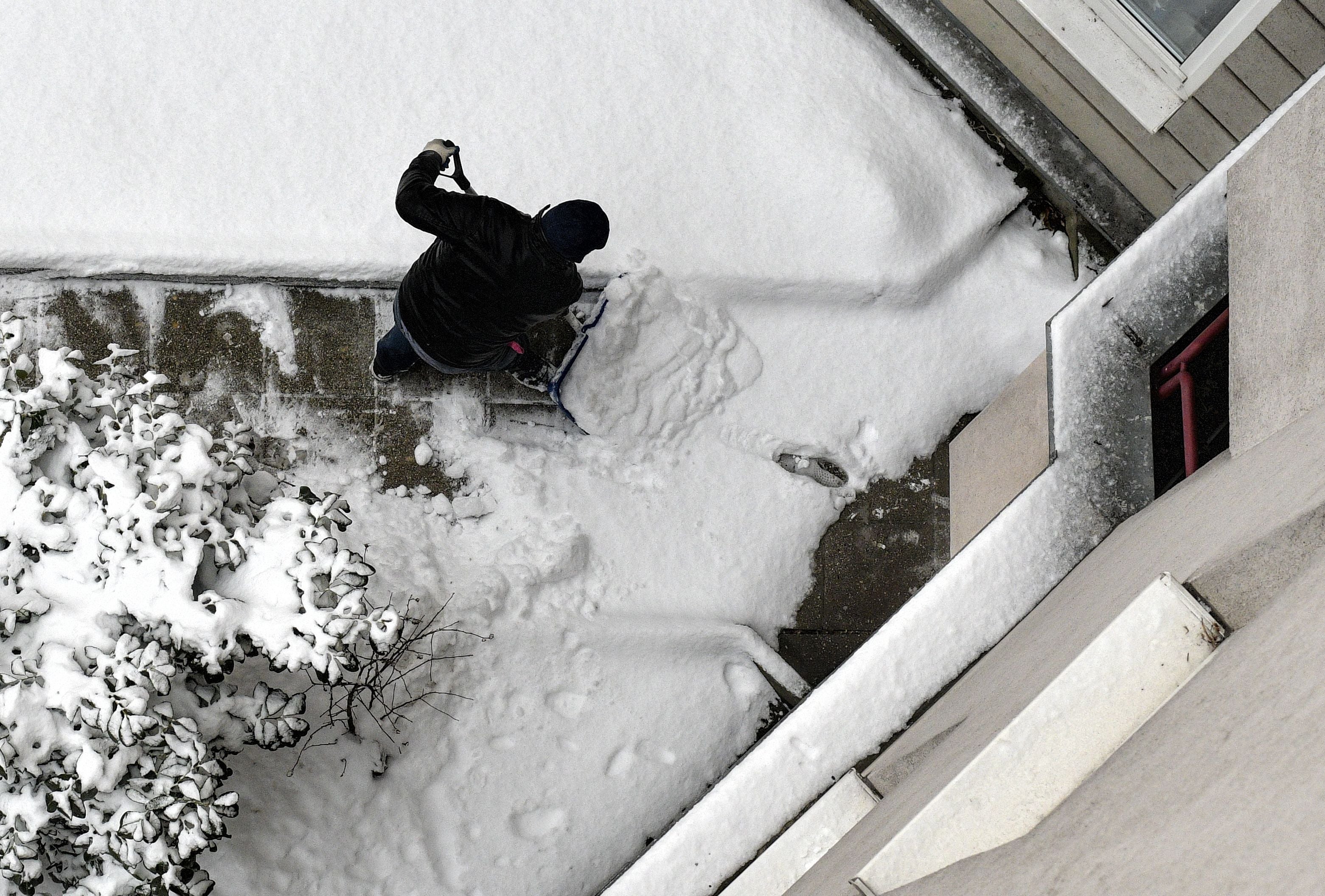 Ein Mann räumt mit einer Schneeschaufel Schnee von einem Gehweg in einer Wohnanlage.