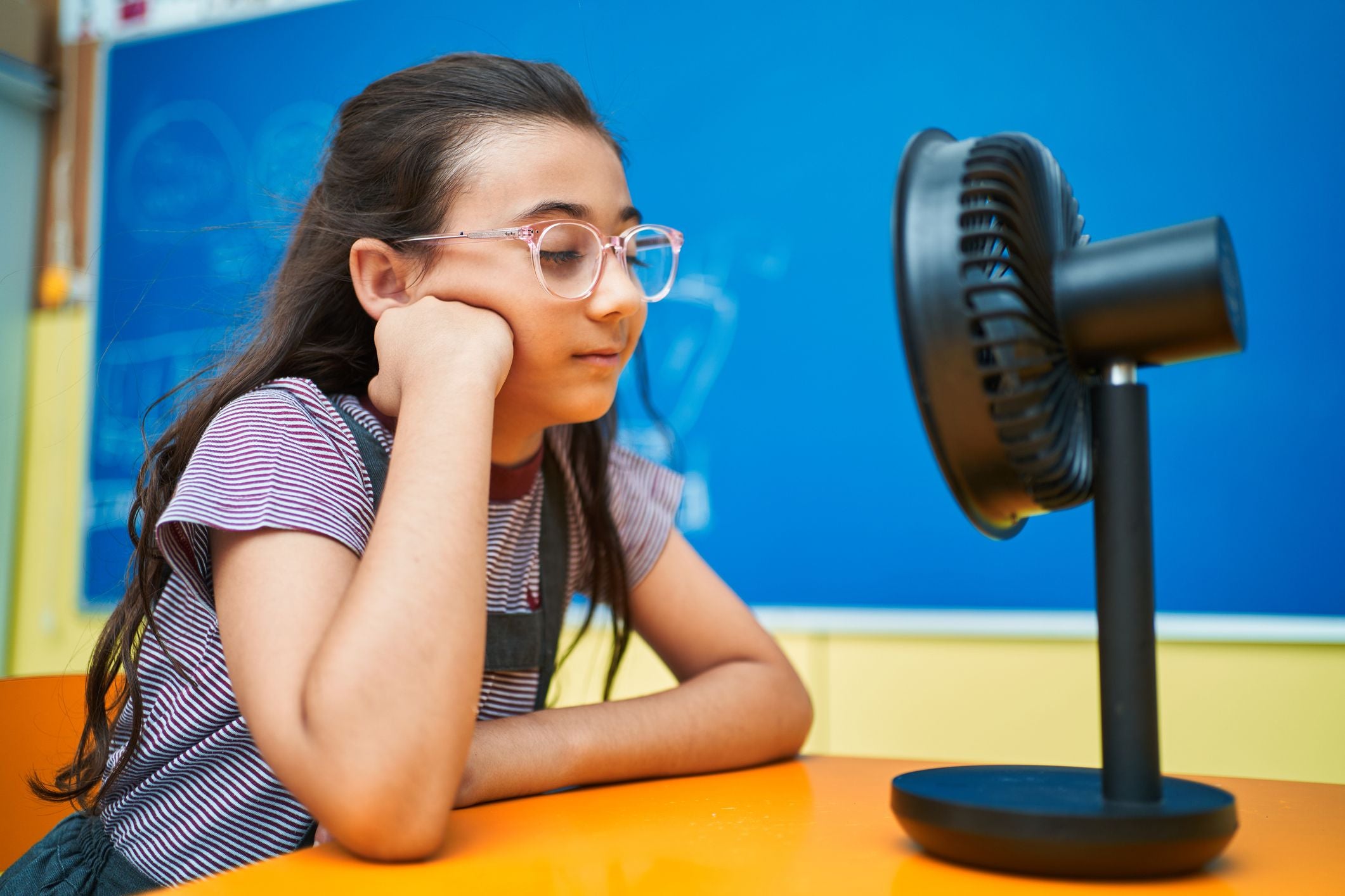 Mädchen sitzt vor einem Ventilator und kühlt sich an einem heißen Tag im Klassenzimmer ab.