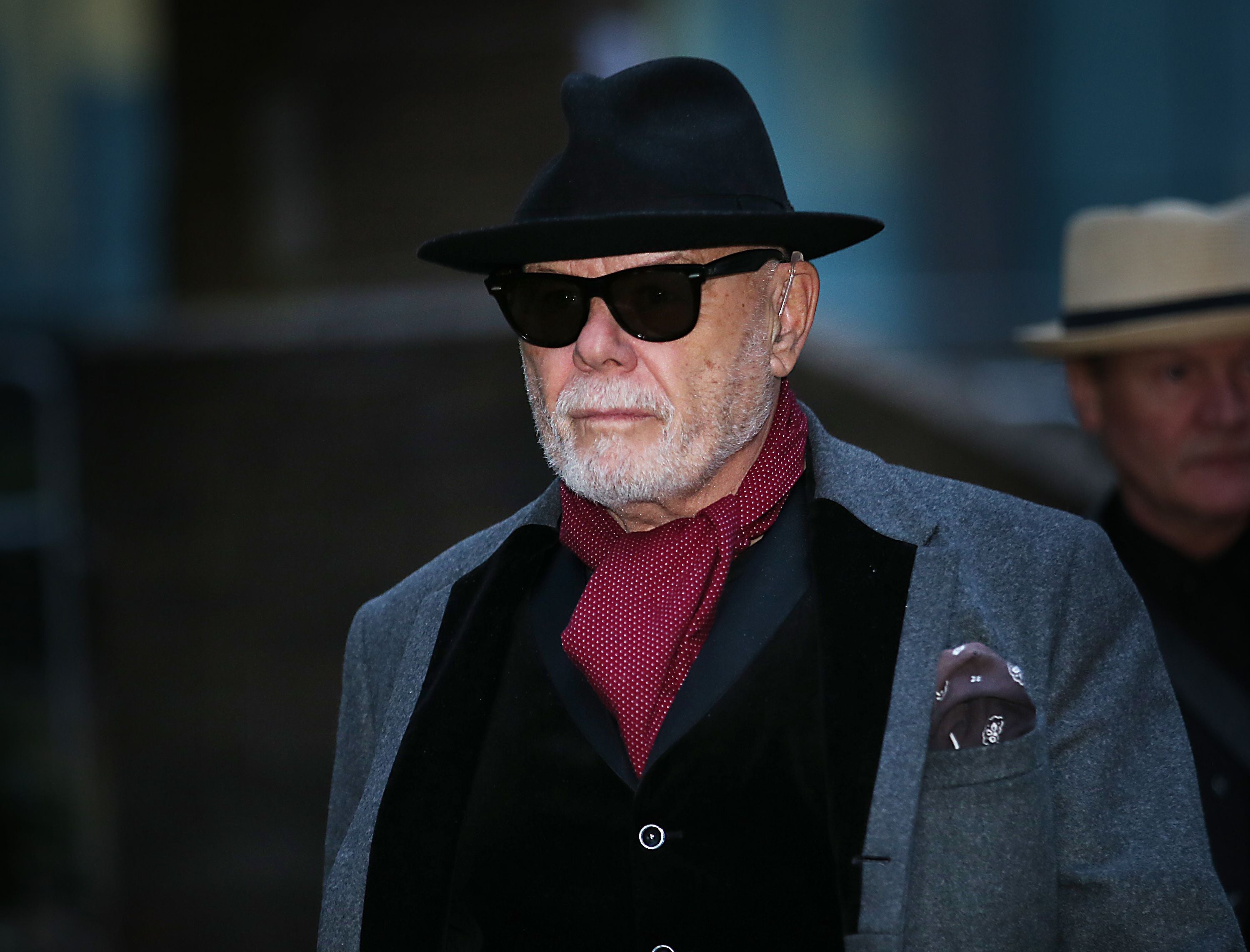 Älterer Mann mit grauem Bart, Hut, Sonnenbrille und rotem Schal auf einer Straße.
