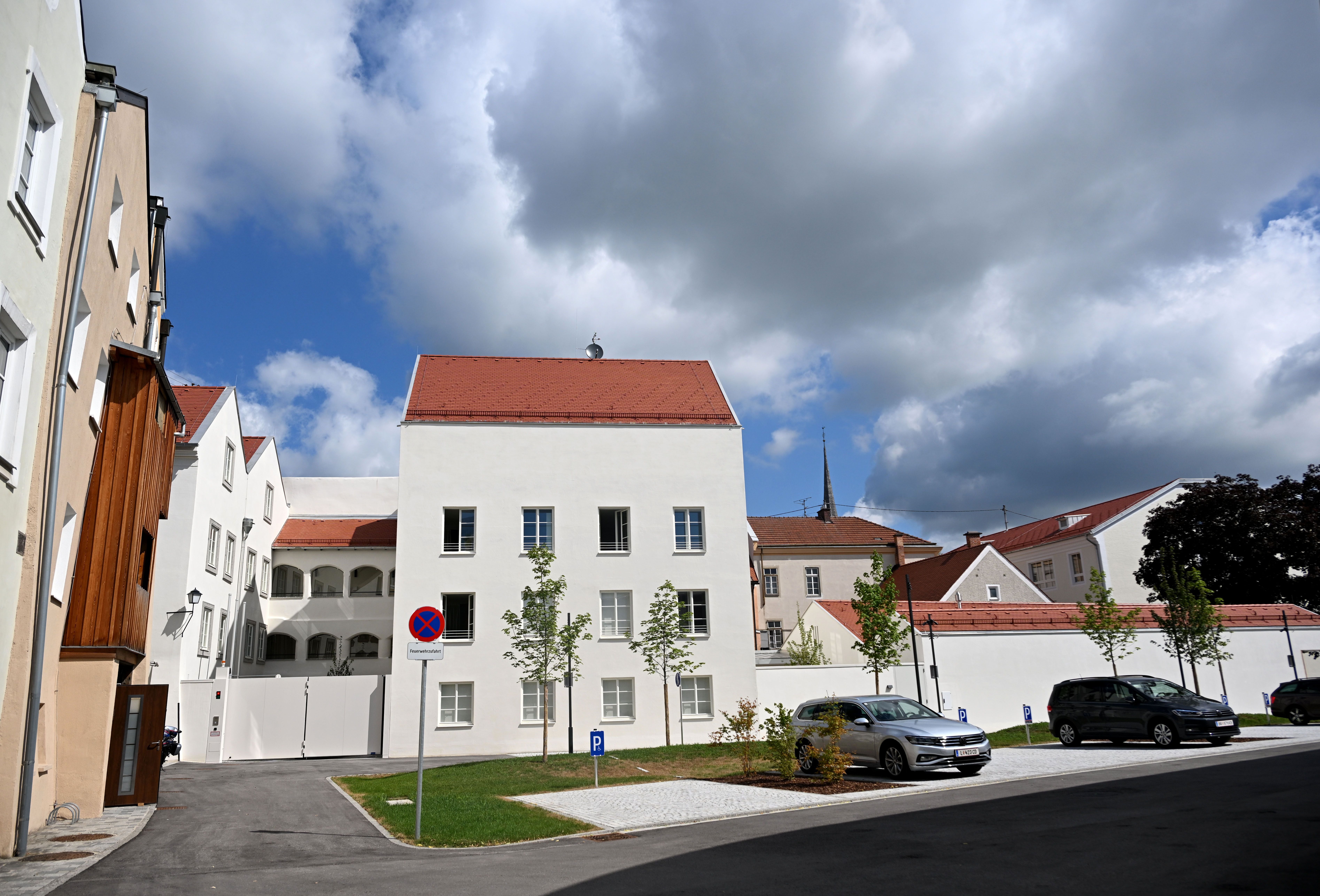 Die Rückseite eines renovierten Gebäudes mit Parkplatz, Bäumen und mehreren Autos unter Wolkenhimmel.