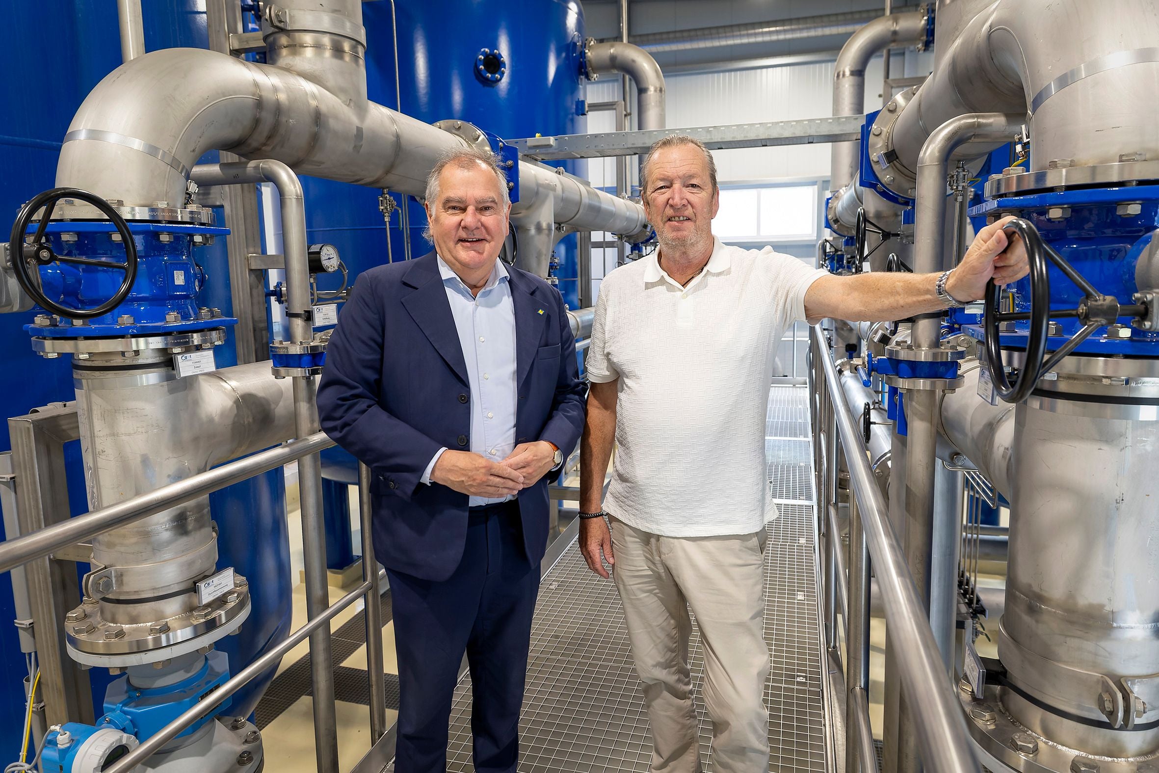 Zwei Männer stehen in einem modernen Wasserwerk zwischen großen Rohren und Ventilen.