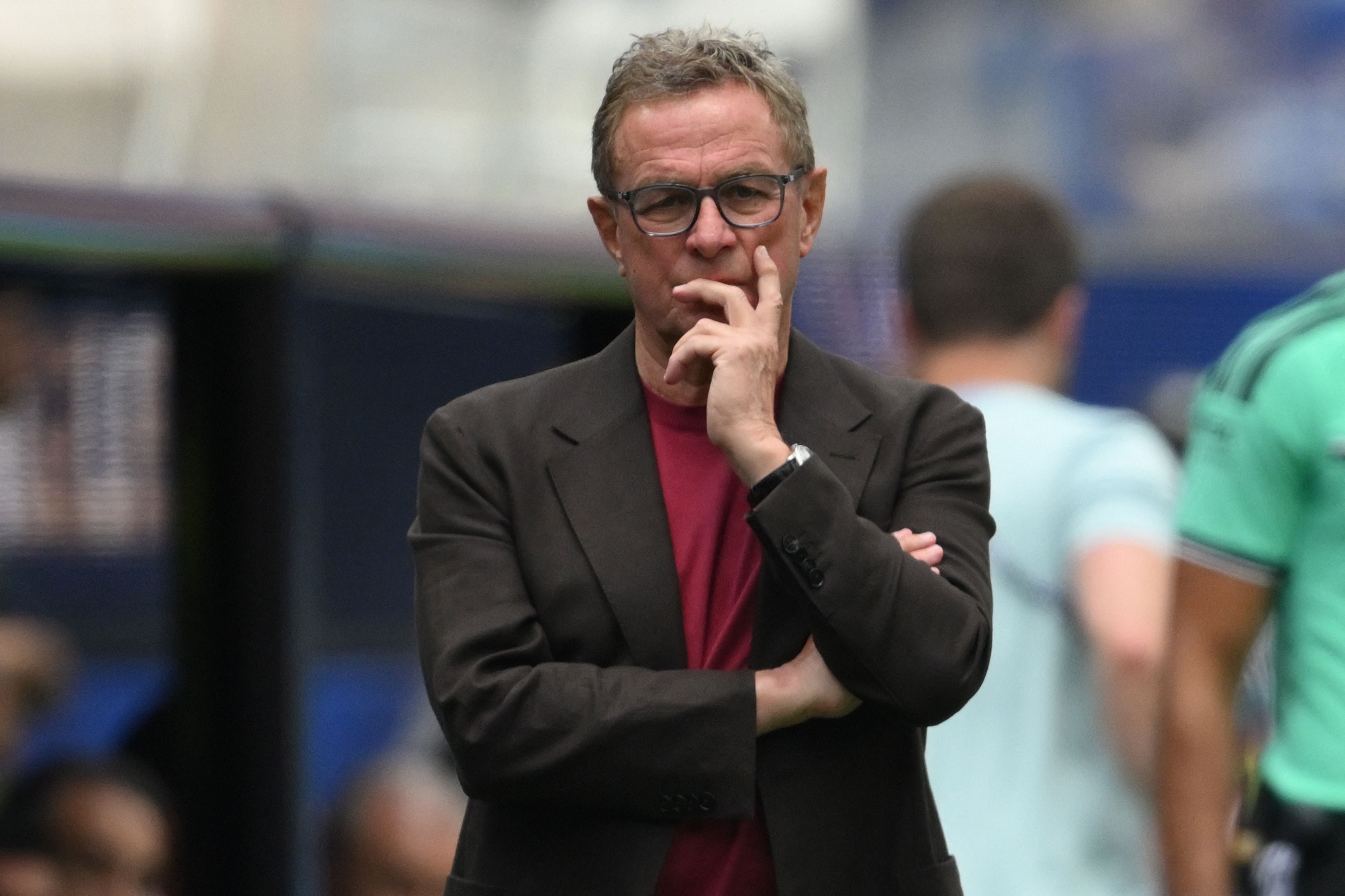 Ein Mann mit Brille und dunklem Sakko steht nachdenklich am Spielfeldrand eines Fußballspiels.