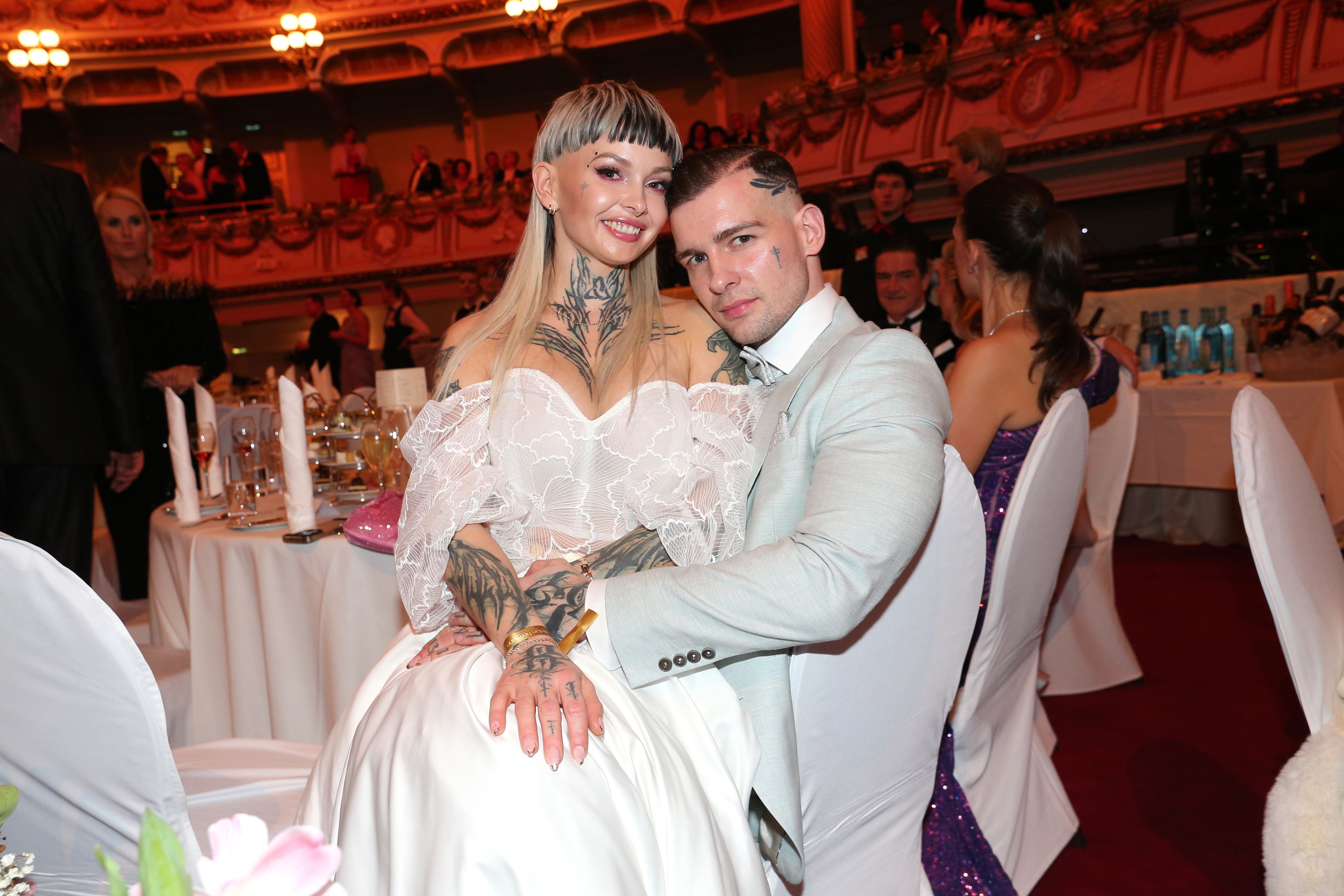 Ein Paar mit auffälligen Tattoos sitzt elegant gekleidet bei einem festlichen Ball.