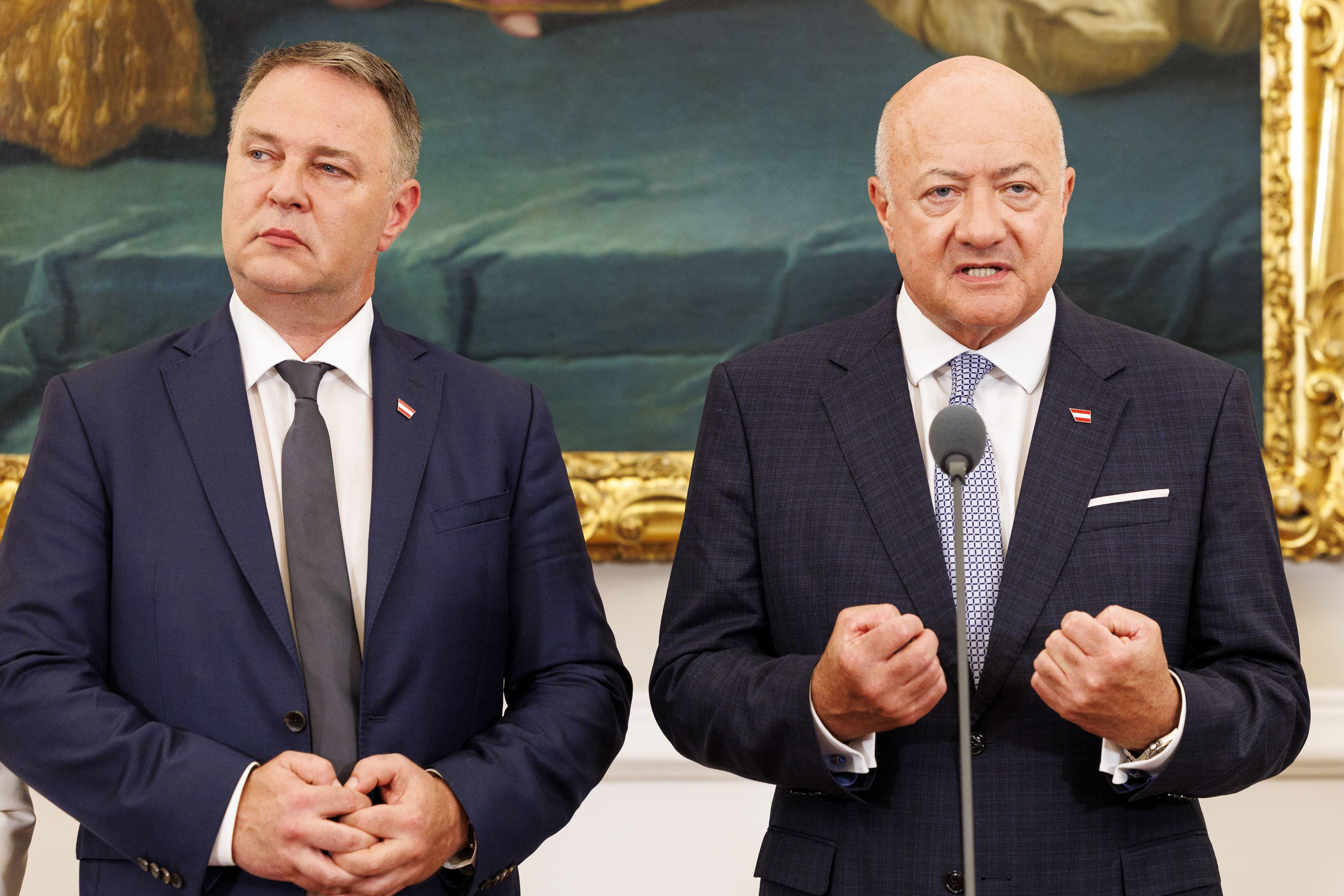 Zwei Männer im Anzug sprechen bei einer Pressekonferenz vor einem Gemälde und Mikrofon.