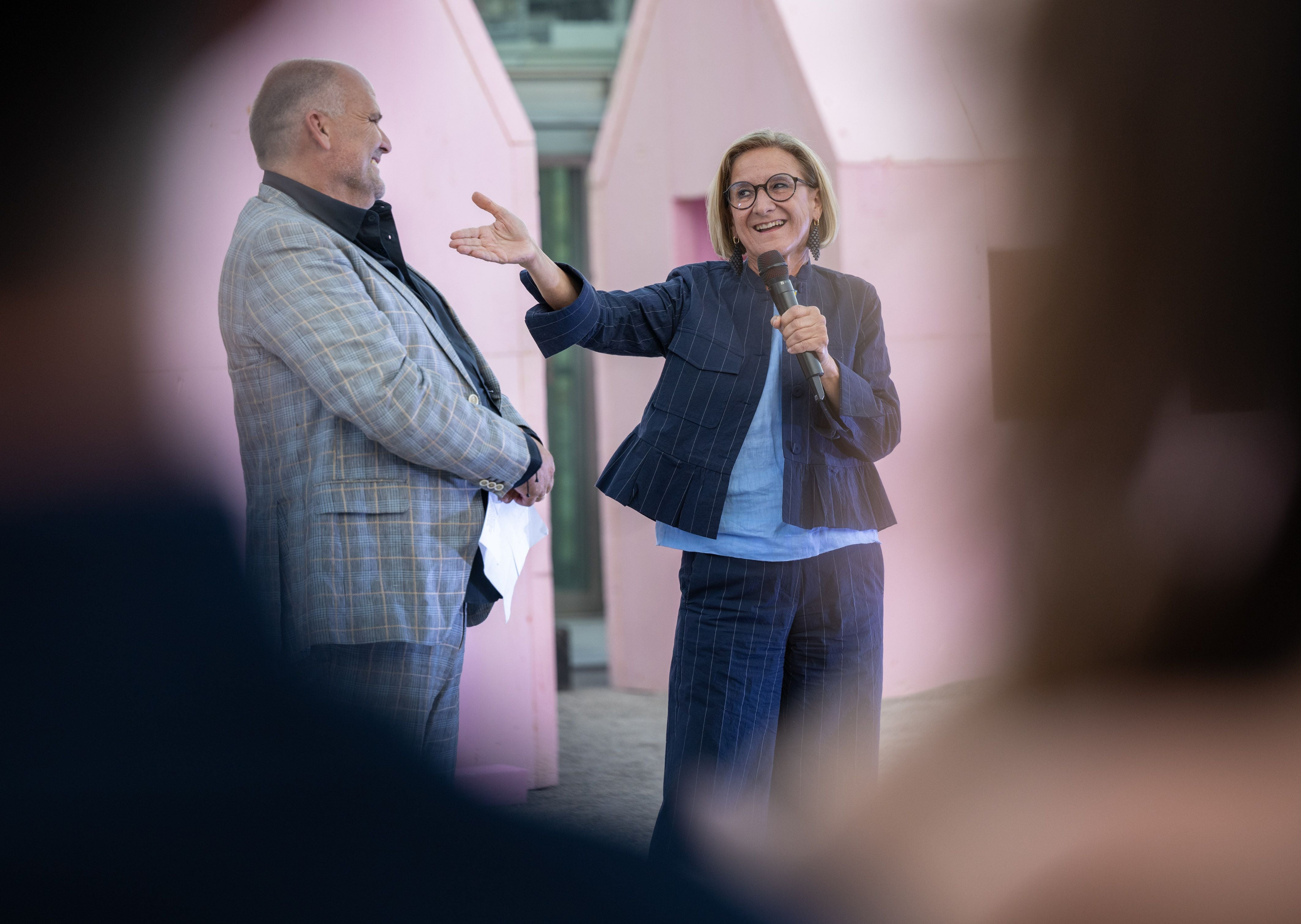 Eine Frau spricht mit Mikrofon neben einem Mann vor rosa Kulisse auf einer Veranstaltung.