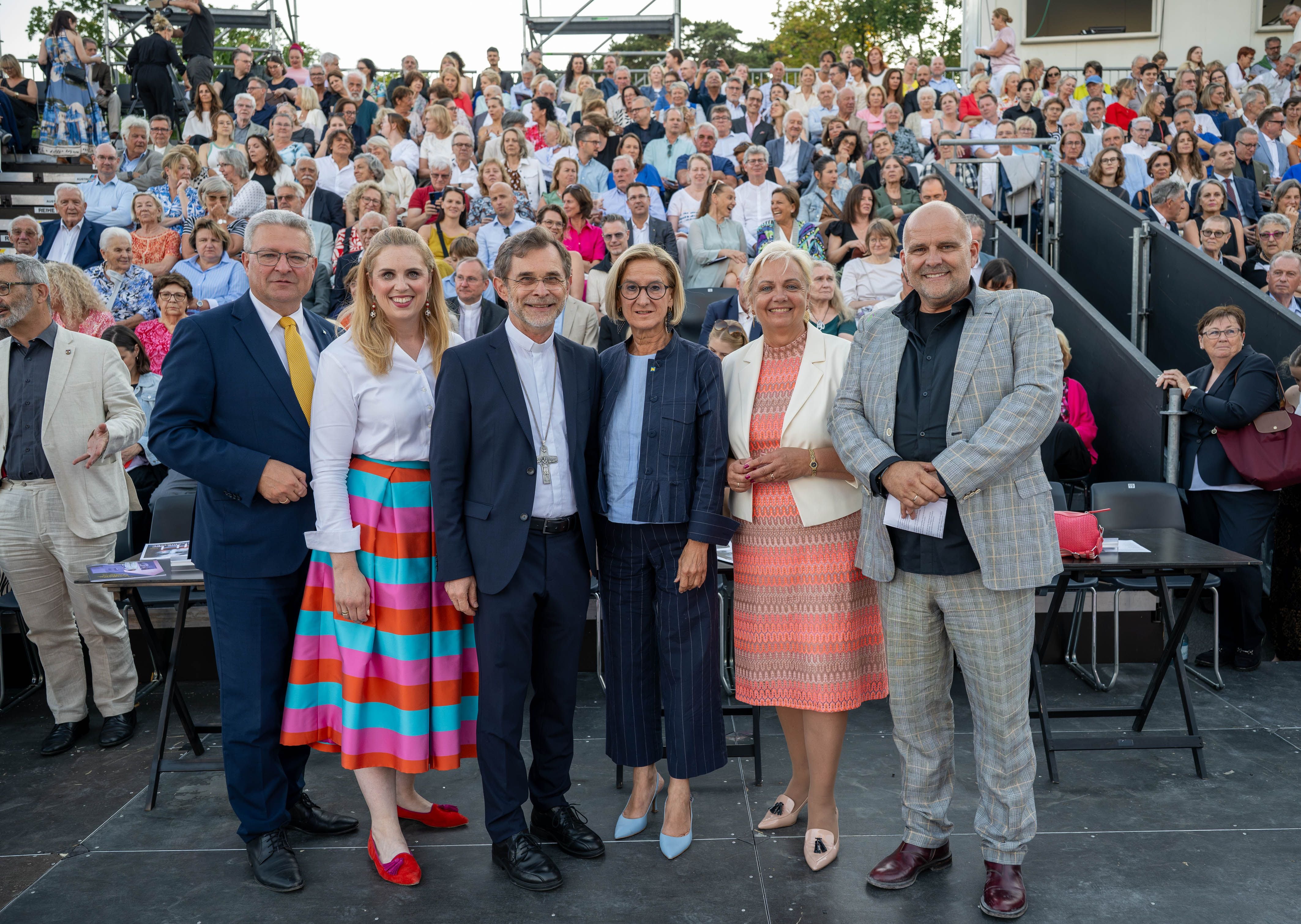 Sechs Personen stehen vor einer großen, sitzenden Menschenmenge bei einer Freiluftveranstaltung.