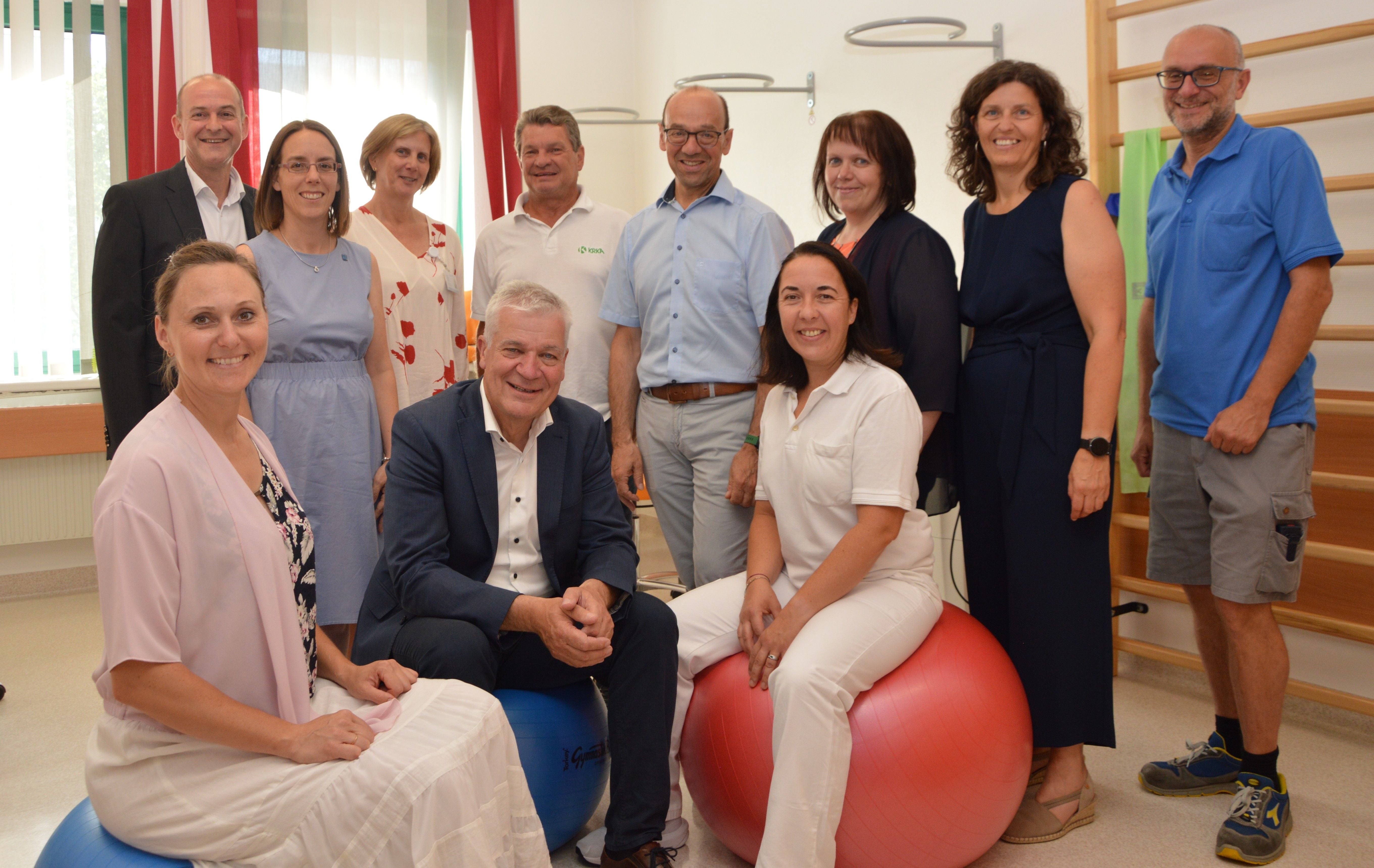 Gruppenfoto von zwölf Erwachsenen in einem hellen Raum, zwei sitzen auf Gymnastikbällen.
