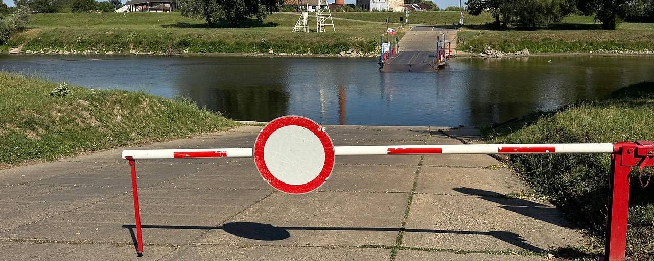 Geschlossene Schranke blockiert Zufahrt zu einer stehenden Autofähre an einem Flussufer.