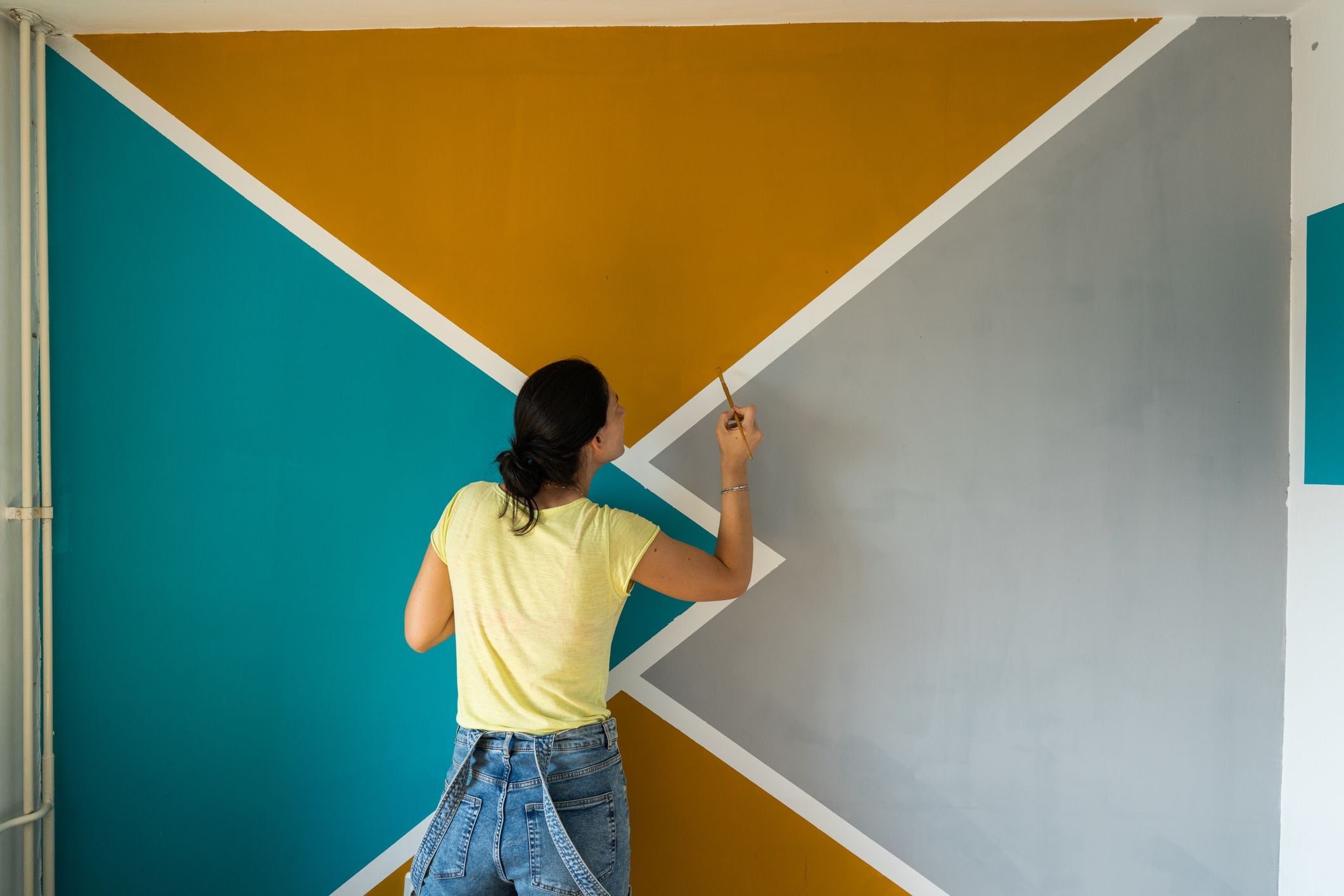 Frau bemalt eine Wand mit geometrischen Mustern in Türkis, Gelb und Grau.