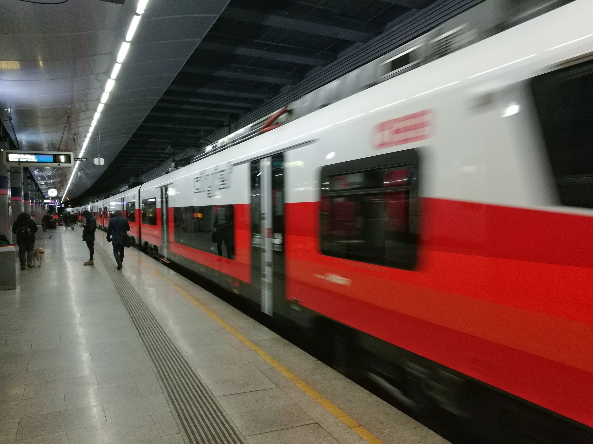Ein rot-weißer Zug fährt in einen modernen, gut beleuchteten Bahnhof ein; wenige Menschen am Bahnsteig.
