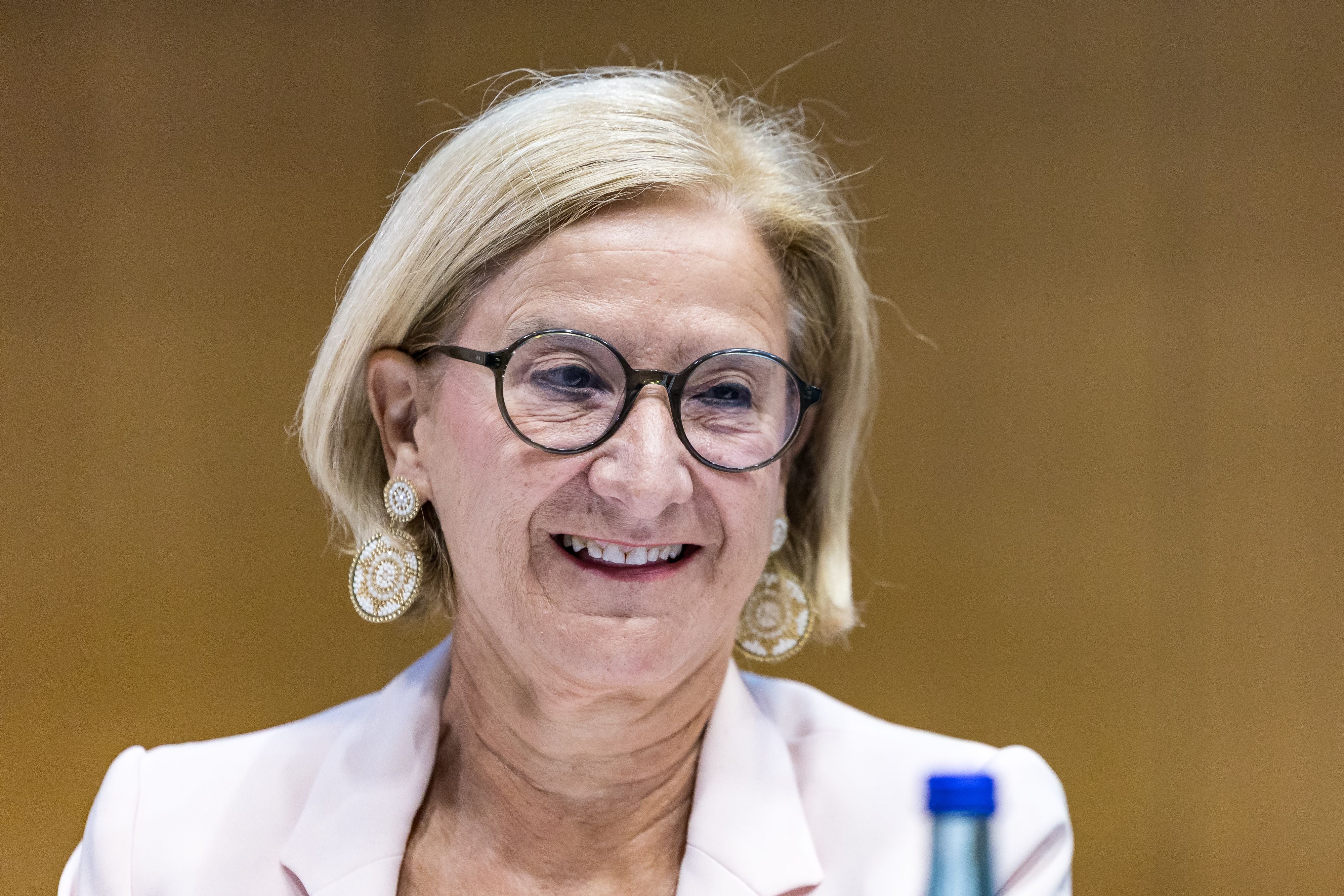 Frau mit Brille und hellblondem Haar lächelt, trägt helle Kleidung und auffällige Ohrringe.