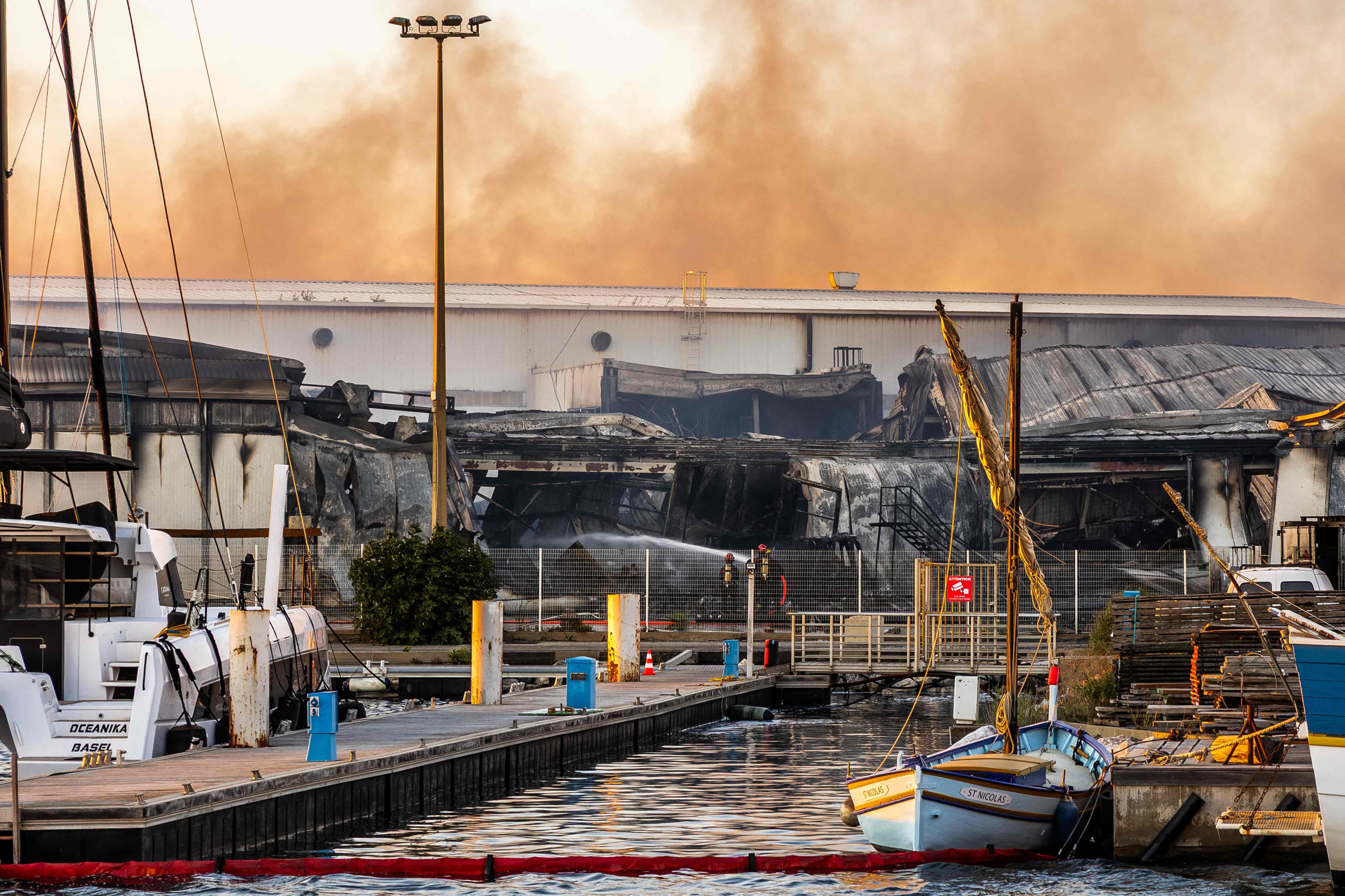 Feuerwehrleute löschen Brand in einer stark beschädigten Werfthalle nahe einem Yachthafen, Rauch steigt auf.