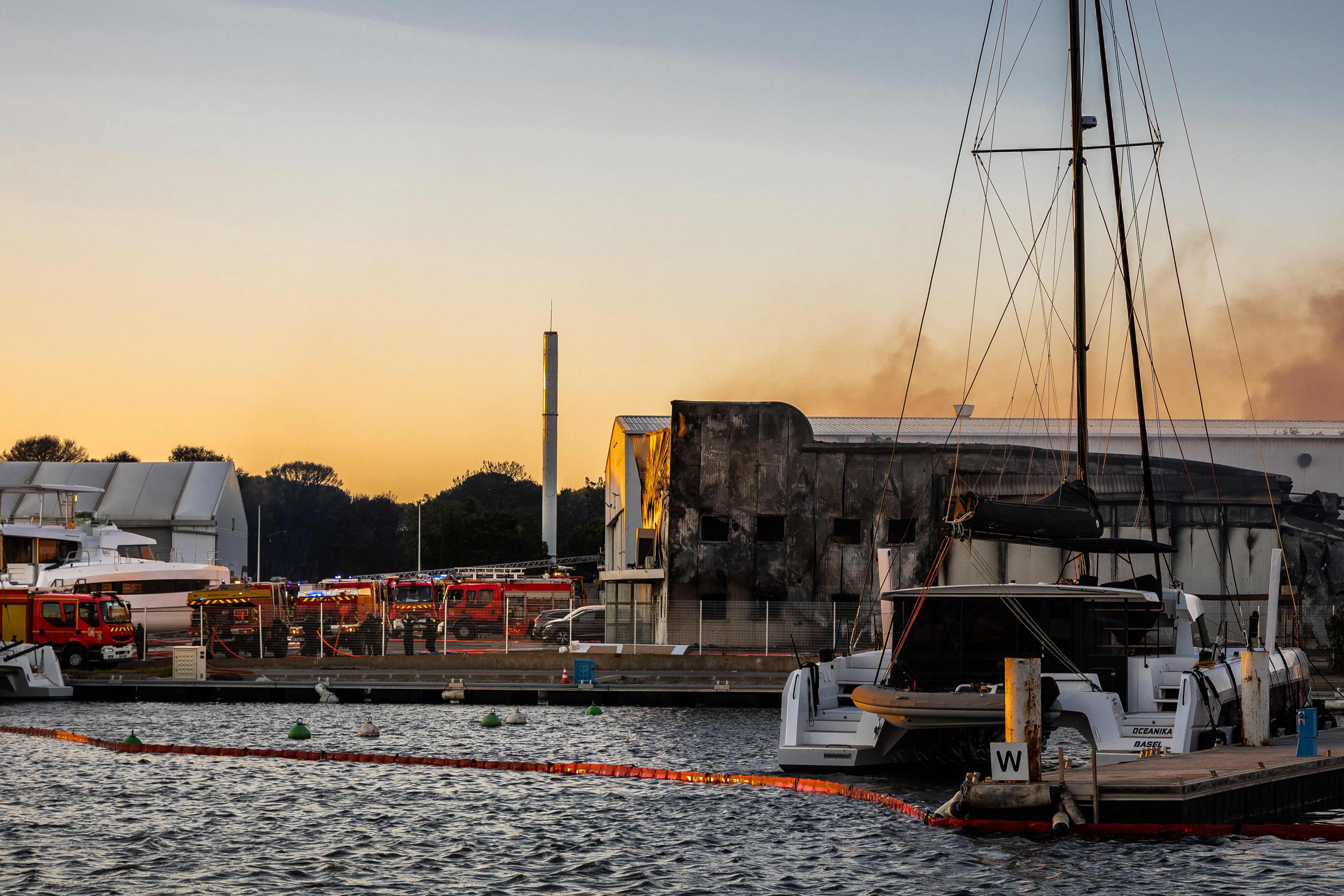 Feuerwehrfahrzeuge neben einem ausgebrannten Gebäude am Hafen, Rauch steigt in den Abendhimmel.