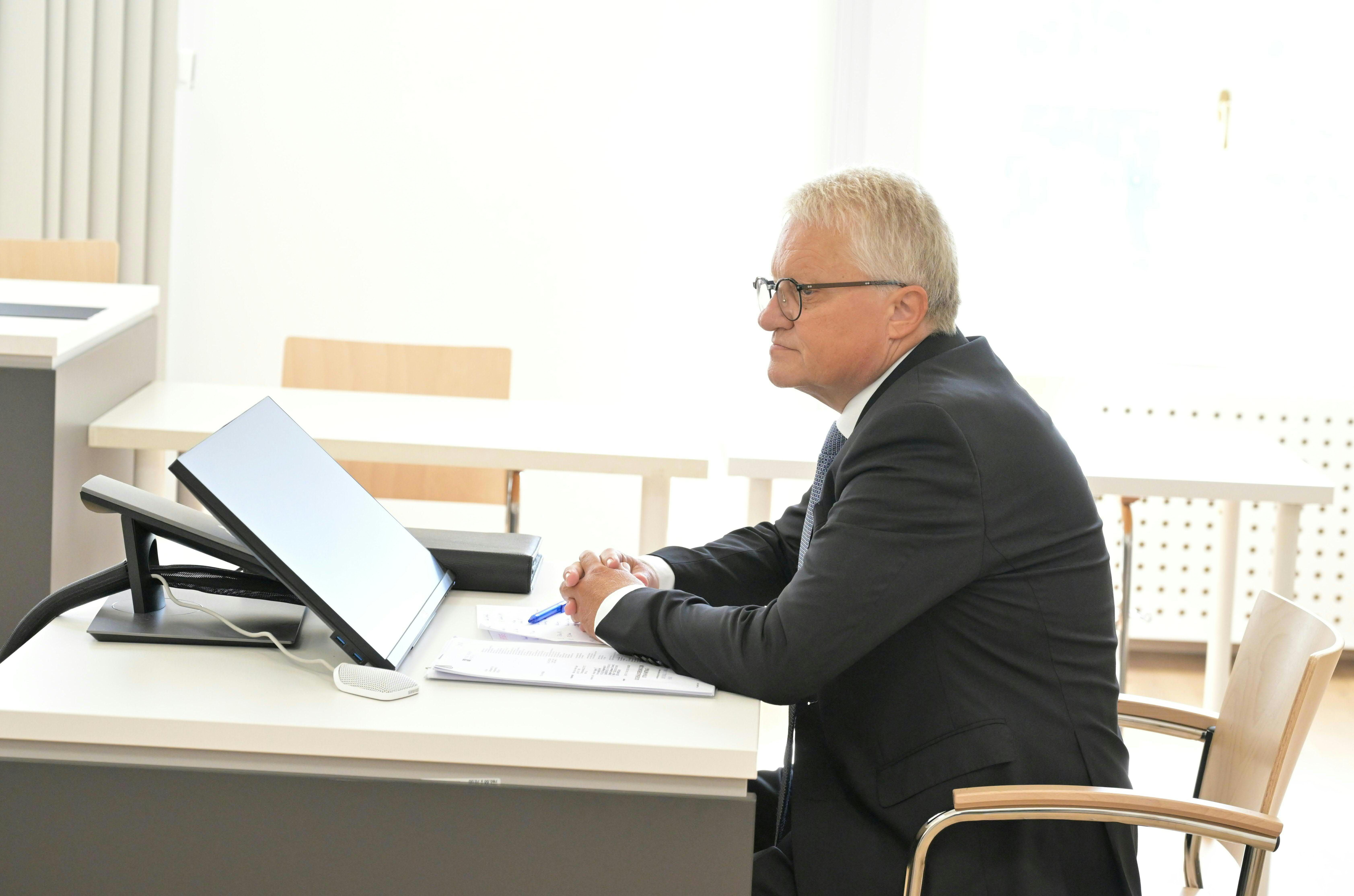 Ein Mann im Anzug sitzt an einem Schreibtisch mit Monitor und schaut nach vorne.