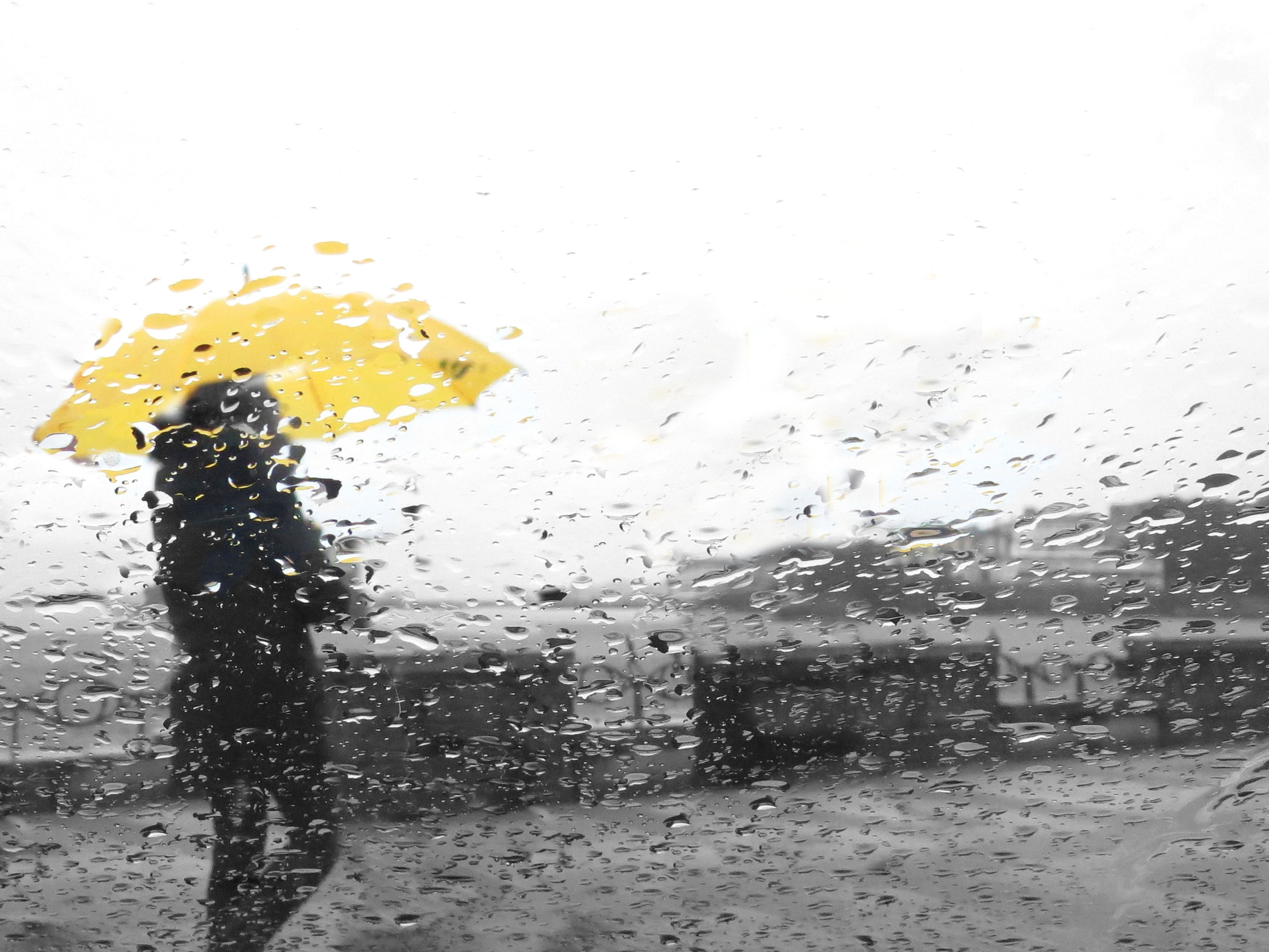 Person mit gelbem Regenschirm bei Regen, gesehen durch ein fenster mit Regentropfen.