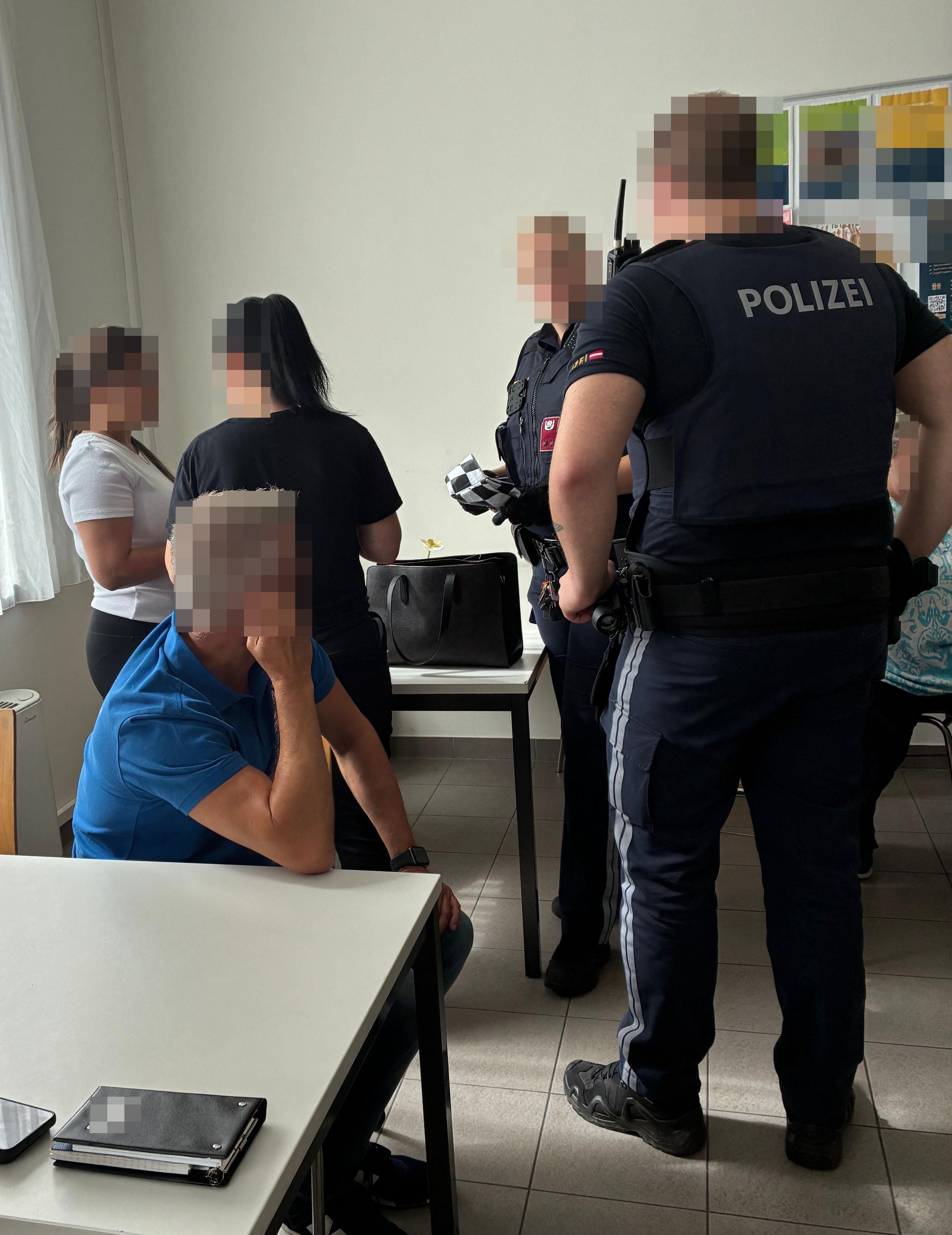 Polizisten und mehrere Personen stehen und sitzen bei einer Kontrolle in einem Zimmer.
