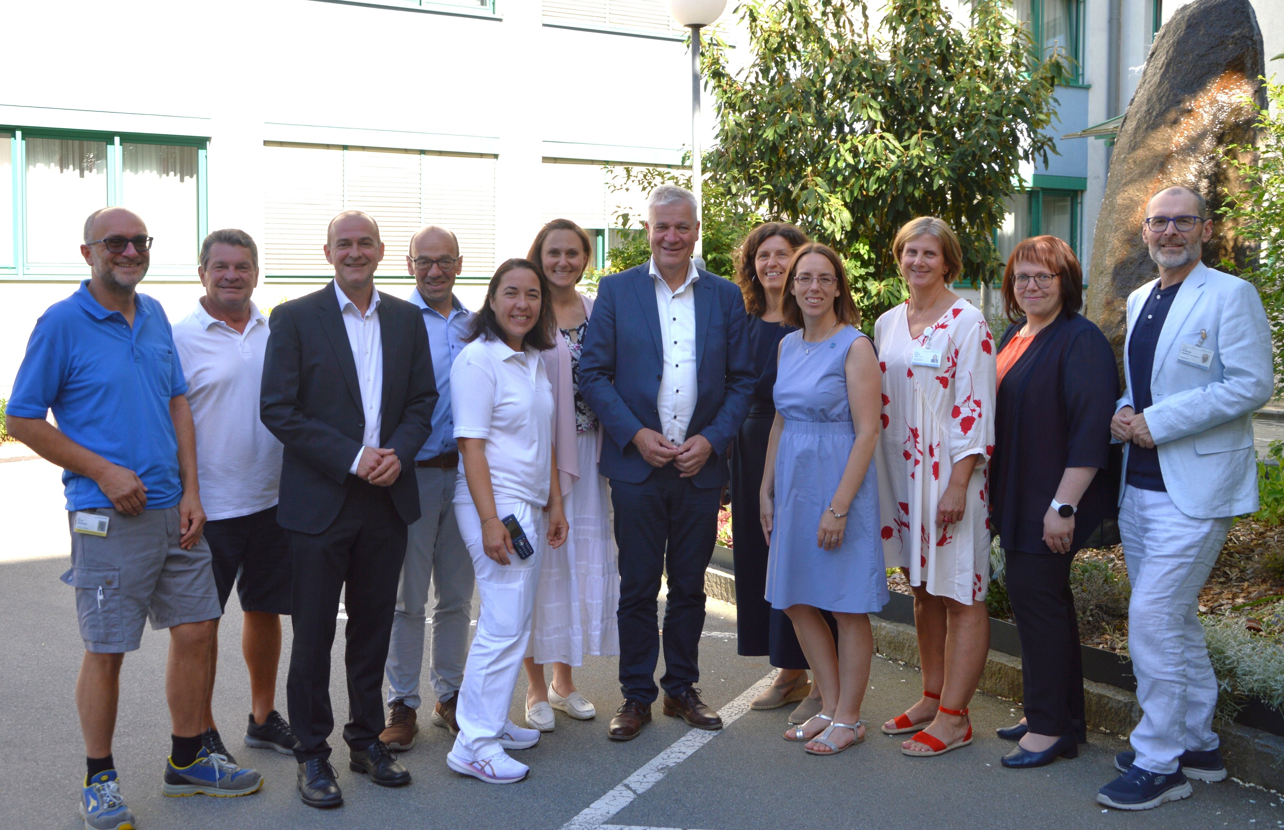 Gruppenfoto von 13 Personen im Freien vor einem Gebäude bei sonnigem Wetter.