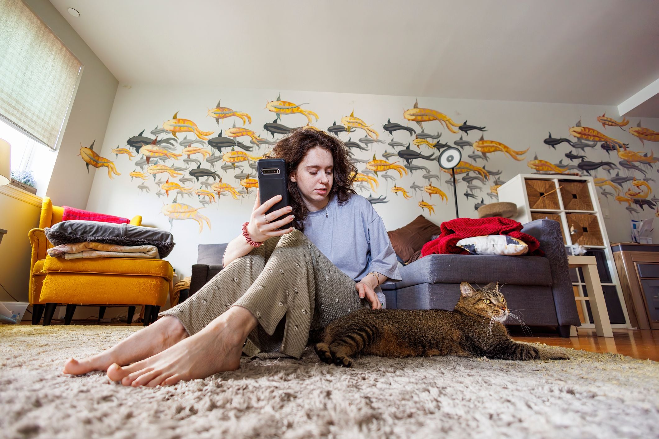 Frau sitzt mit Smartphone auf Teppich und streichelt ihre Katze im Wohnzimmer mit bunter Wandmalerei.