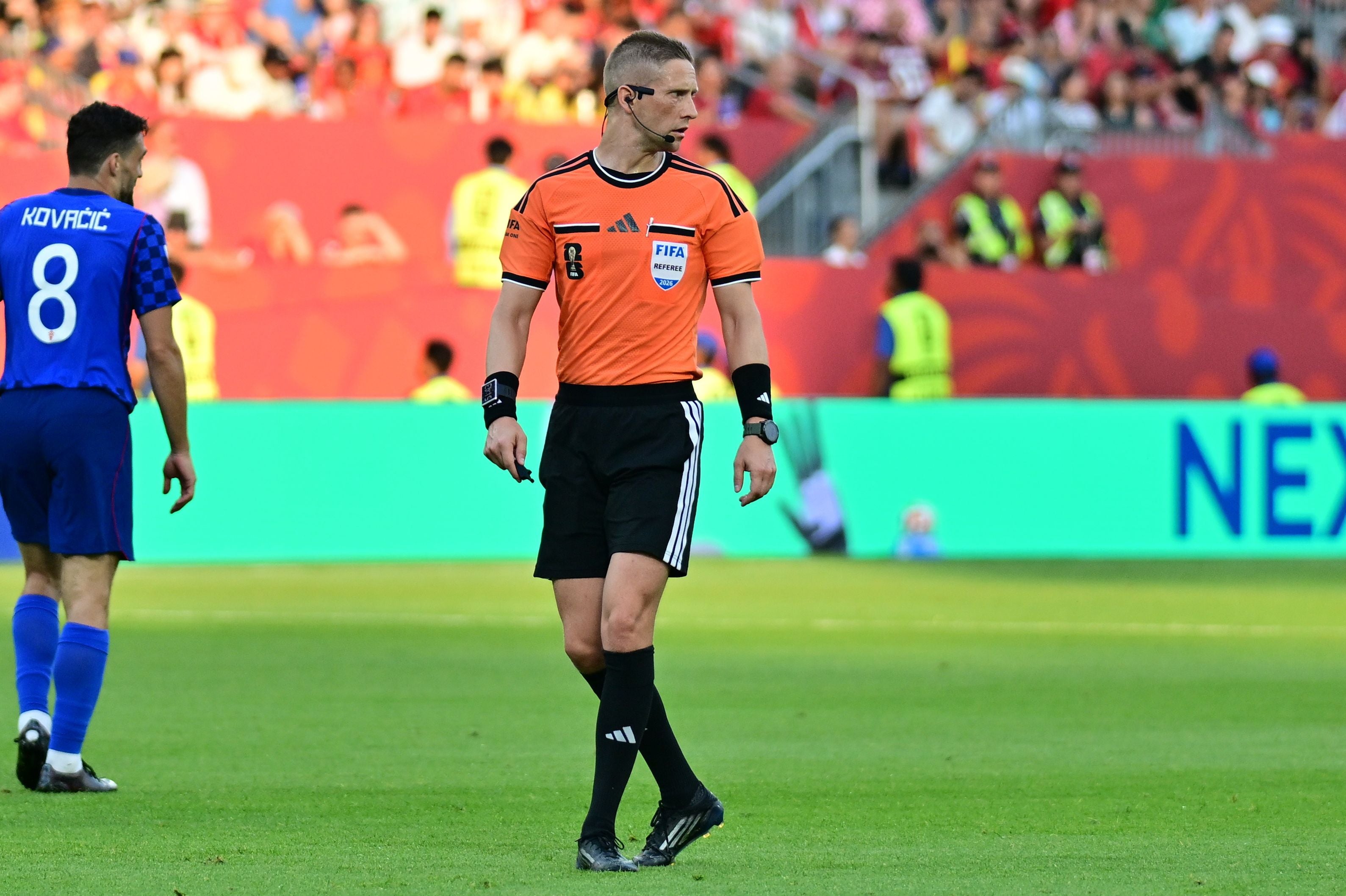 Schiedsrichter in orangem Trikot steht auf dem Fußballfeld, Spieler und Zuschauer im Hintergrund.